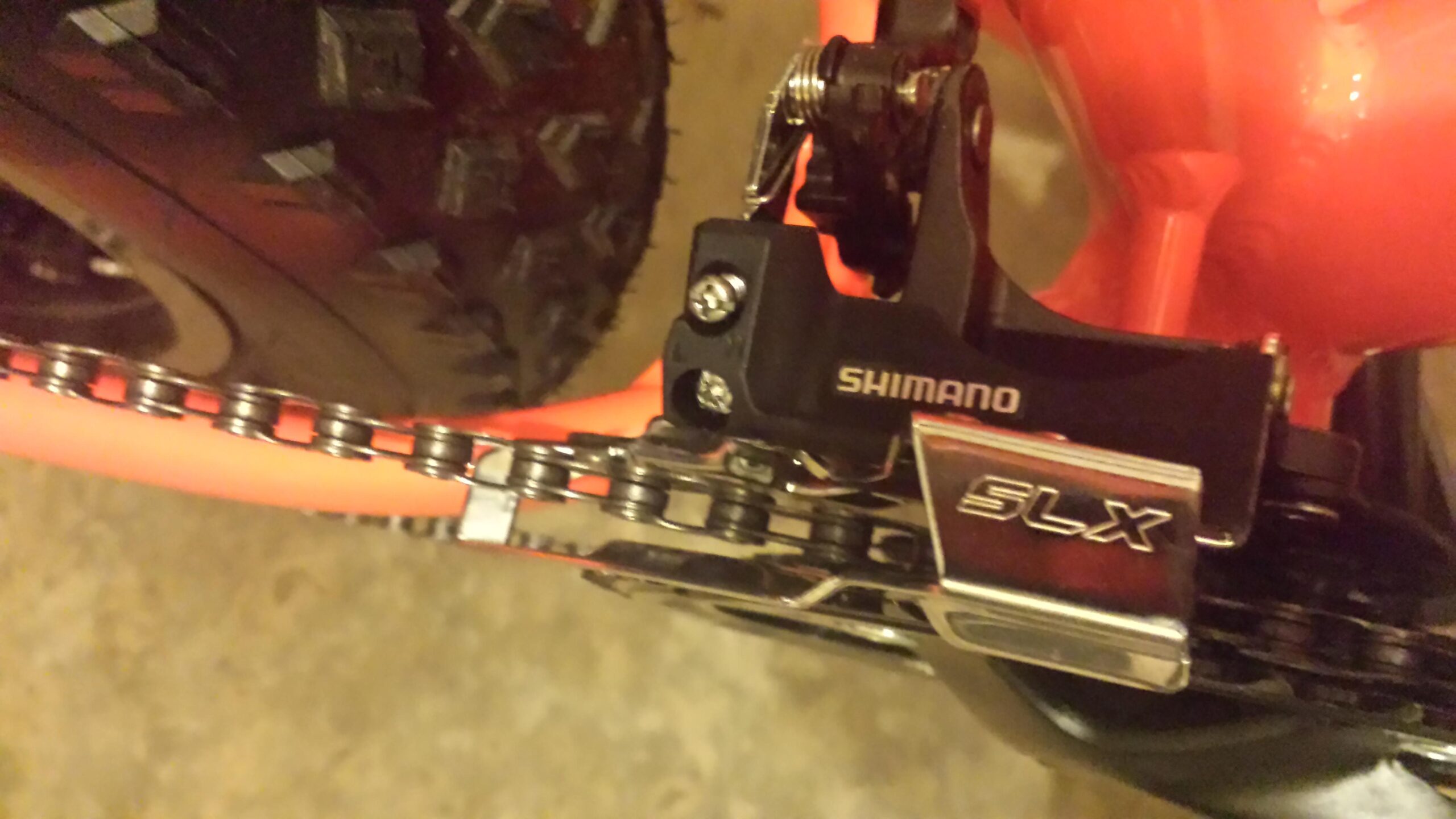 Motobecane FB4 Comp: Close-up view of a Shimano SLX rear derailleur mounted on a bicycle frame, with a focus on the chain, gear, and vibrant orange bike components. The image highlights the detailing of the derailleur and chain alignment against a textured background.