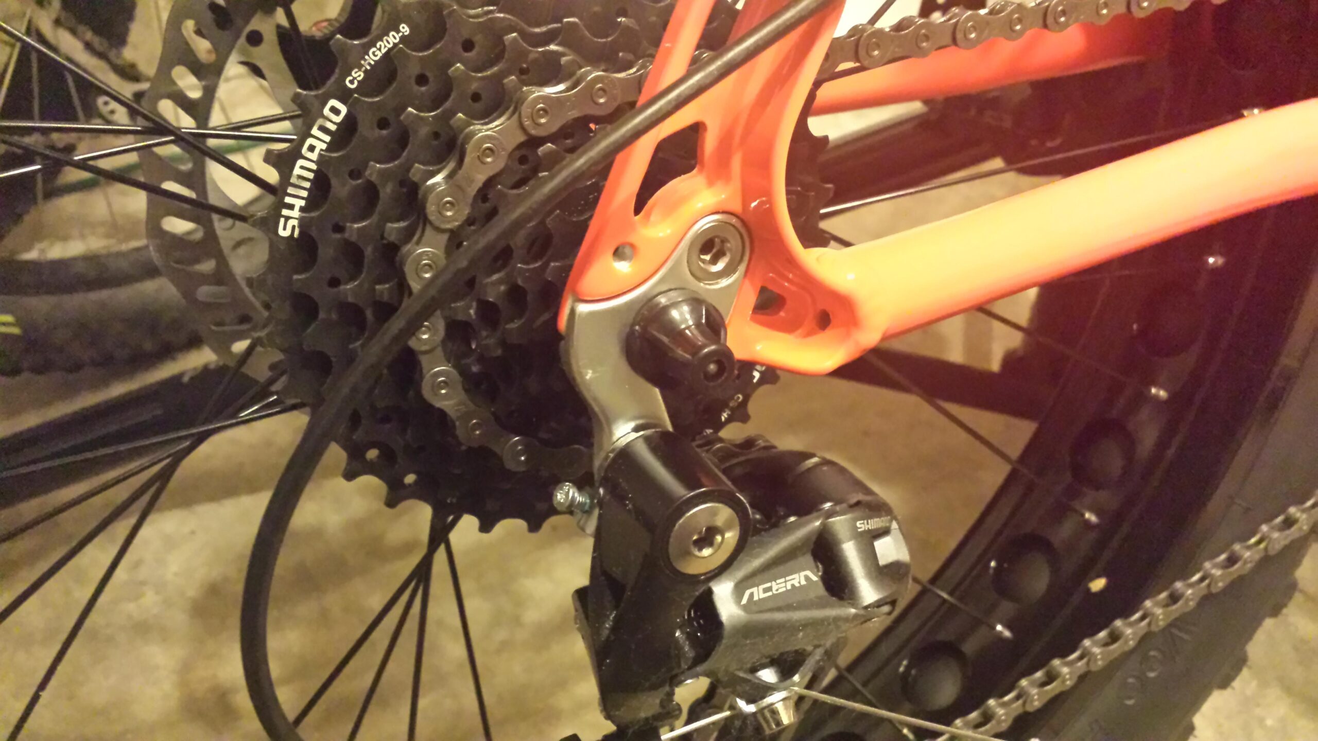Motobecane FB4 Comp: Close-up of a bicycle's rear drivetrain system, featuring a Shimano cassette and derailleur, with an orange frame section visible in the background. The image captures the intricate components of the gear system, including the chain and cables. The setting appears to be indoors, possibly in a garage or workshop.
