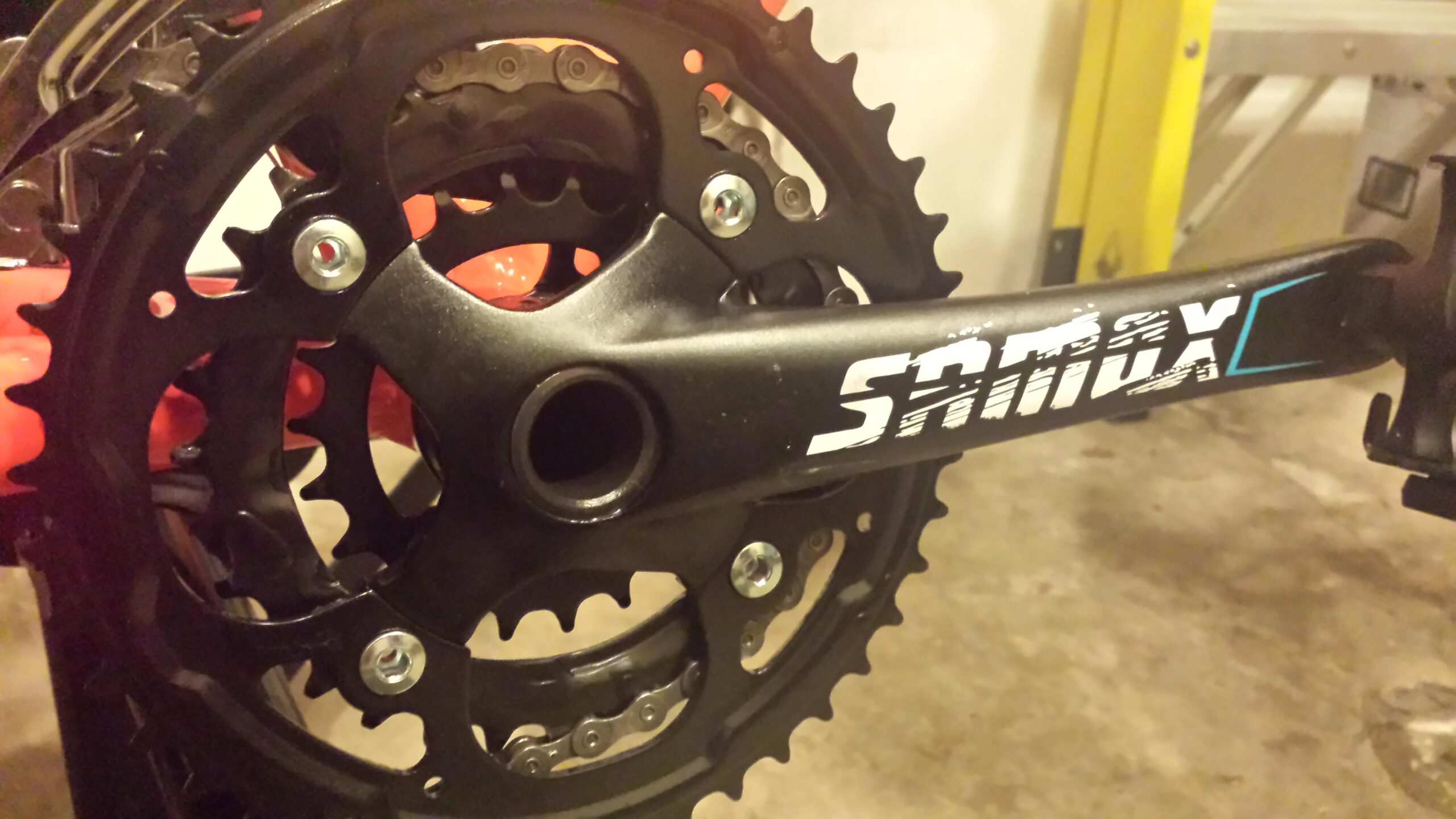 Motobecane FB4 Comp: Close-up of a bicycle crankset showing the black chainring with sharp teeth and the crank arm labeled "Sram X." The background suggests a workshop environment with a partially visible ladder and a concrete floor.