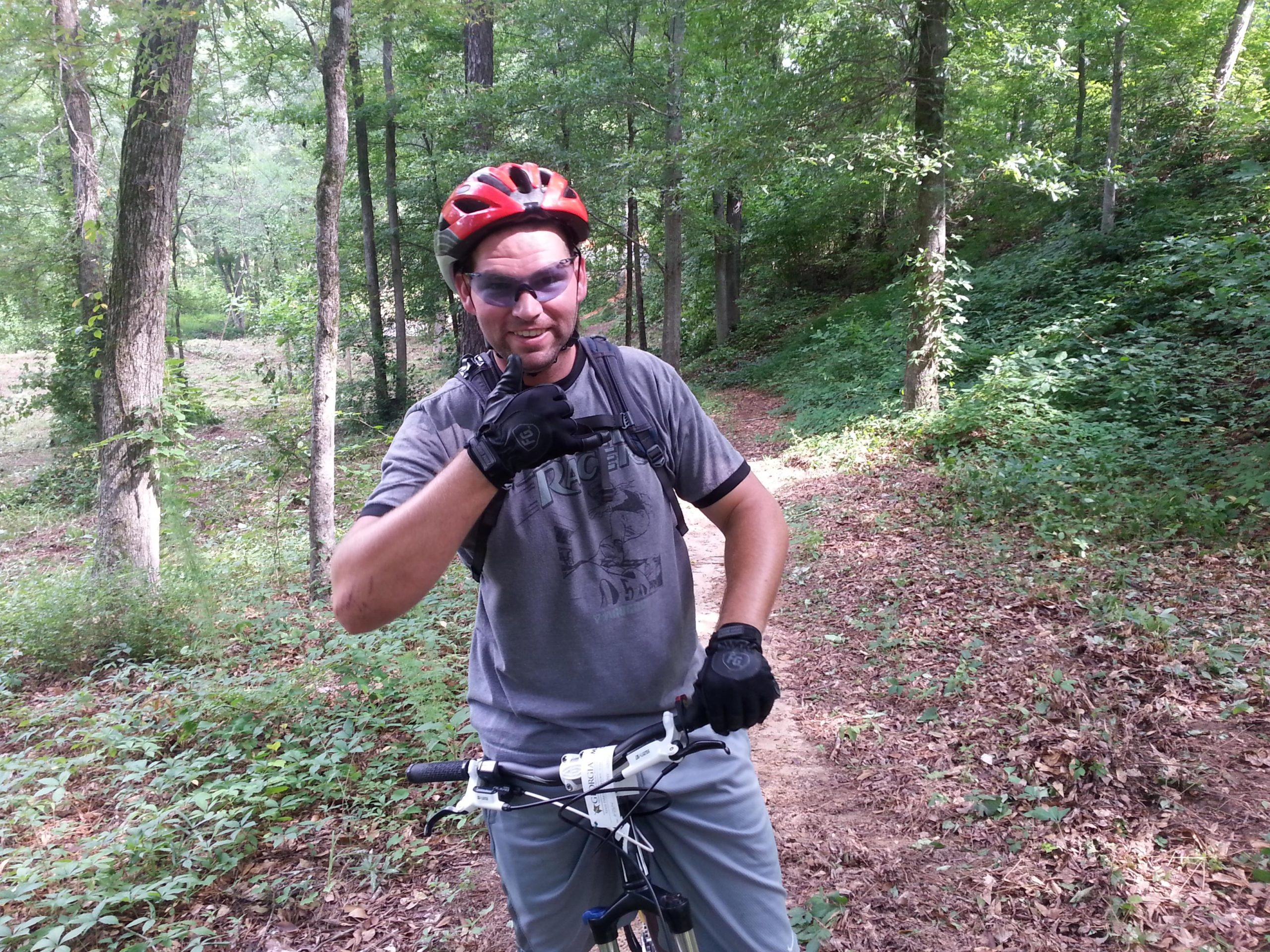 A man wearing a red bicycle helmet and sunglasses stands next to a mountain bike on a dirt trail surrounded by trees. He is making a thumbs-up gesture with one hand while resting the other on the bike handlebar. The scene is set in a green, wooded area with a natural landscape in the background. Fort Yargo State Park mountain bike trail.