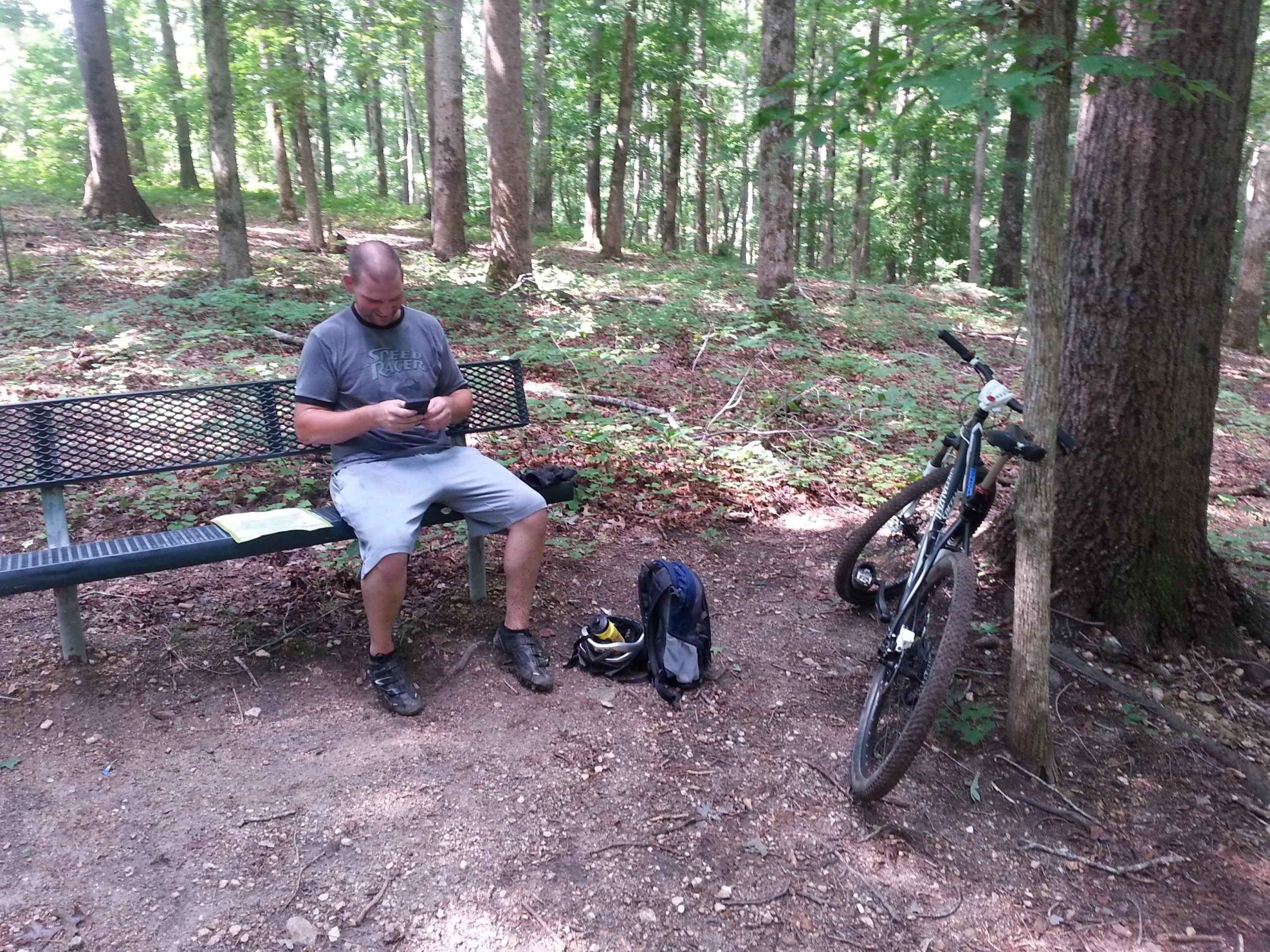 A person sitting on a bench in a forested area, using a mobile phone. Next to them is a backpack and a mountain bike leaning against a tree, surrounded by greenery and sunlight filtering through the trees. Fort Yargo State Park mountain bike trail.