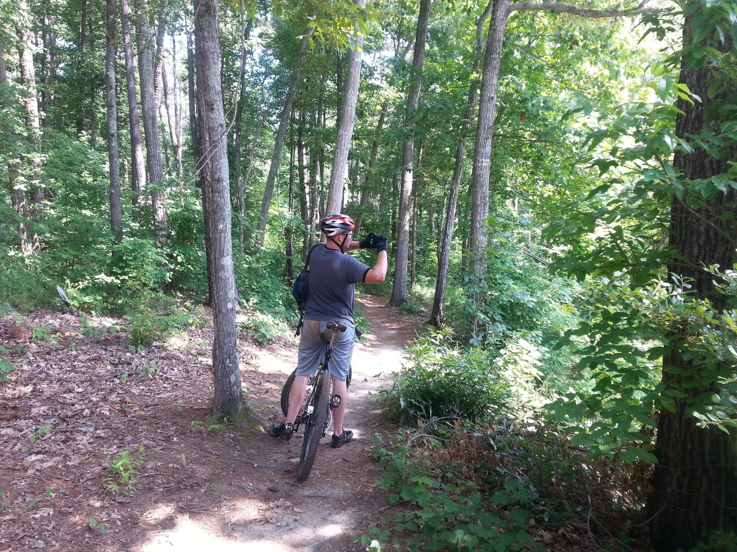 A person wearing a helmet and cycling gear stands beside their mountain bike on a narrow dirt trail in a dense forest. They are looking back down the path, surrounded by lush green trees and underbrush. Fort Yargo State Park mountain bike trail.
