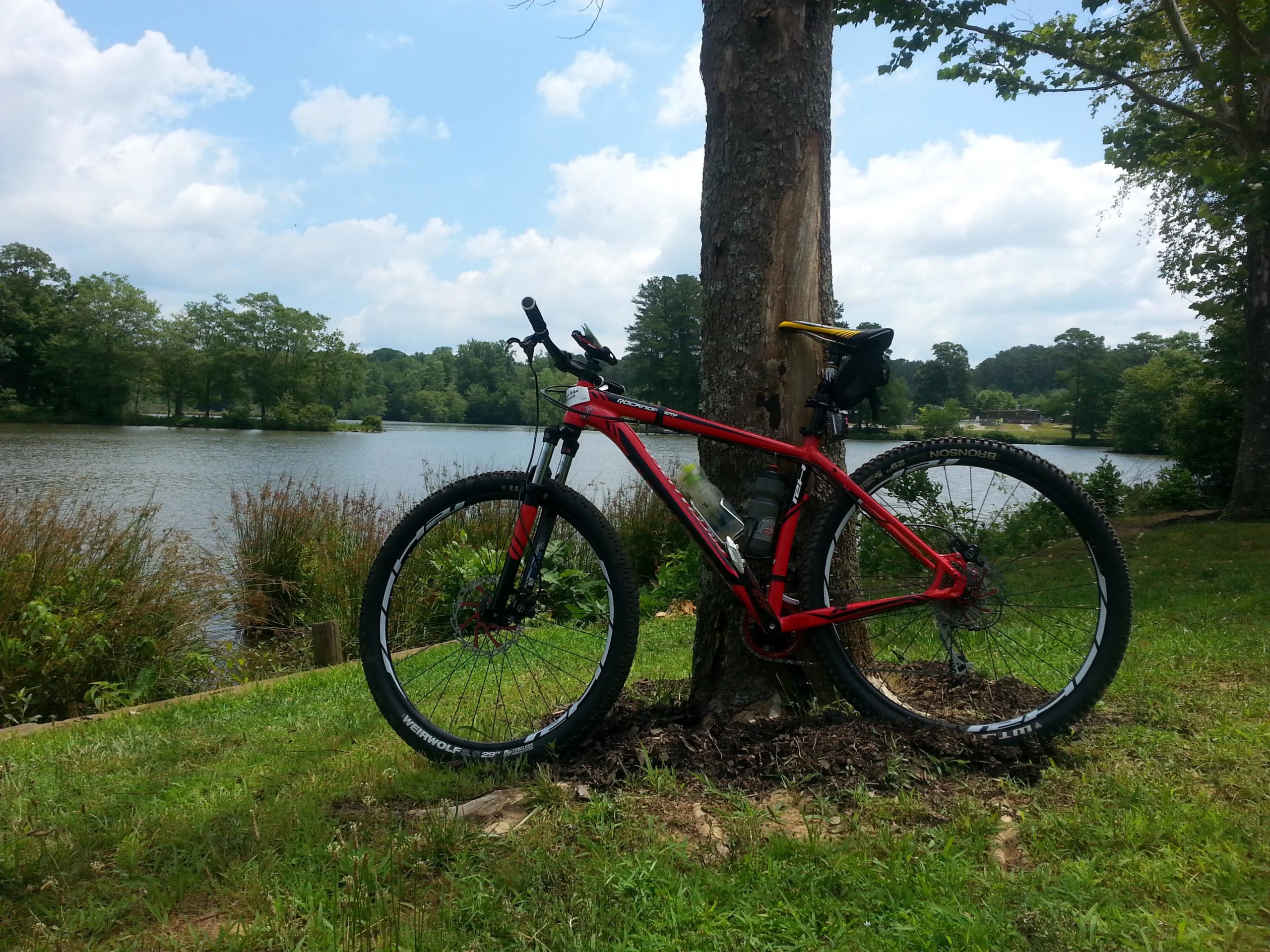 A red mountain bike leaning against a tree in a grassy area near a calm lake, with trees and a cloudy sky in the background. Fort Yargo State Park mountain bike trail.