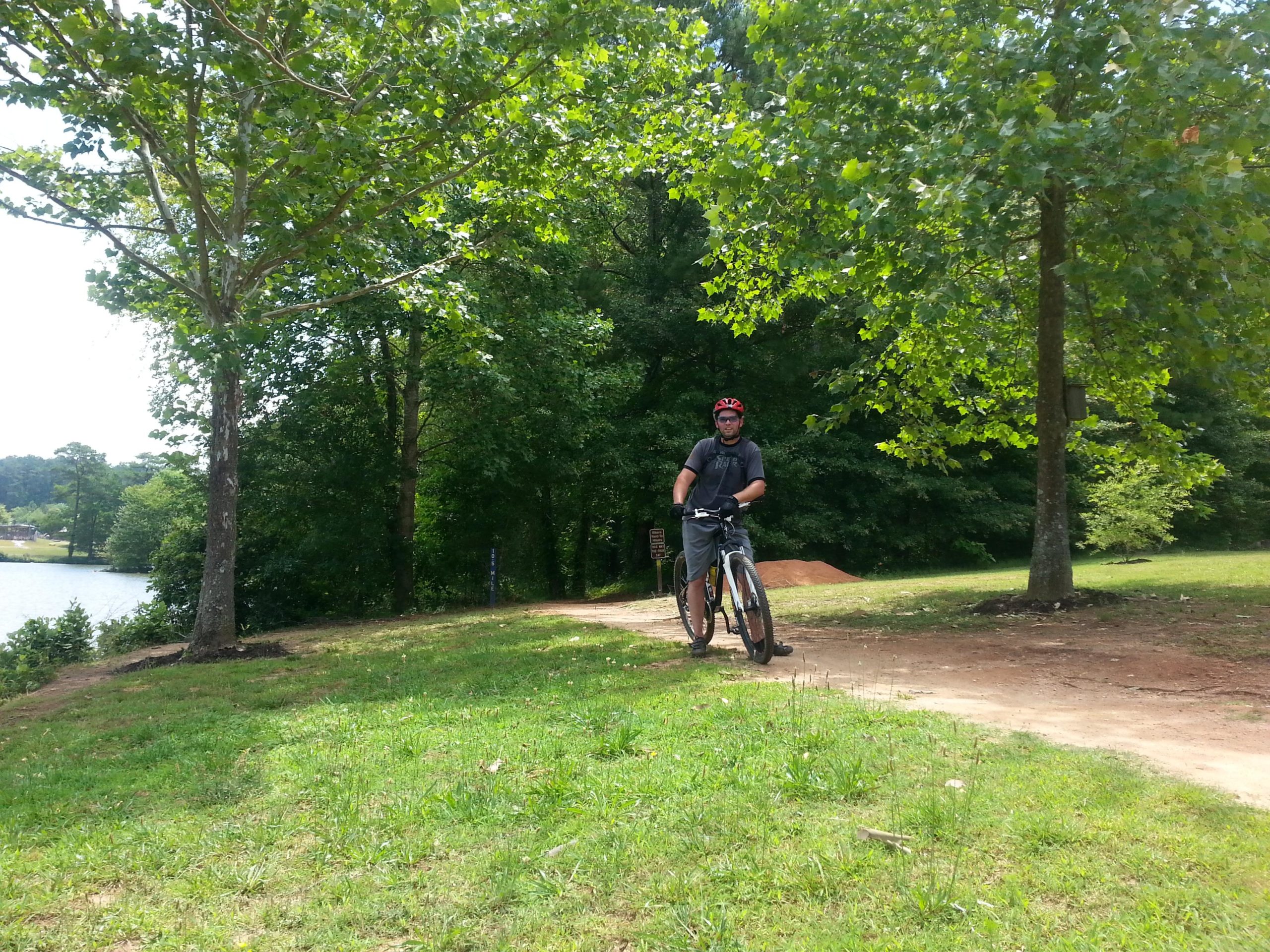 A person wearing a helmet and gloves stands next to a mountain bike on a dirt path beside a lake, surrounded by green trees and grass on a sunny day. Fort Yargo State Park mountain bike trail.