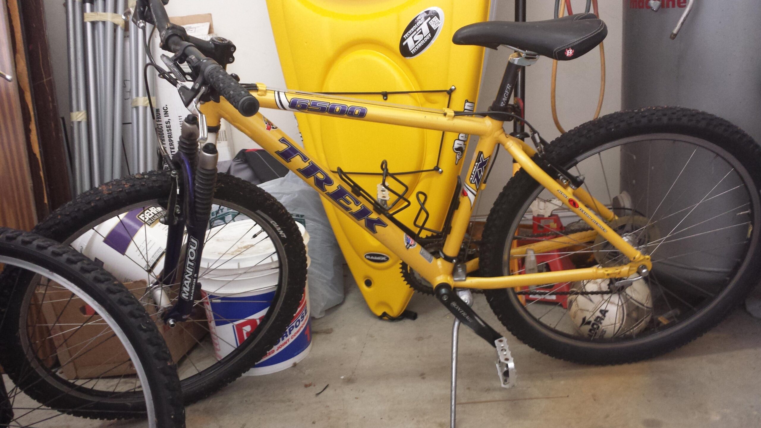 A yellow Trek mountain bike leaning against items in a garage, with wide tires and front suspension visible. In the background, a yellow kayak and various storage containers are partially visible.