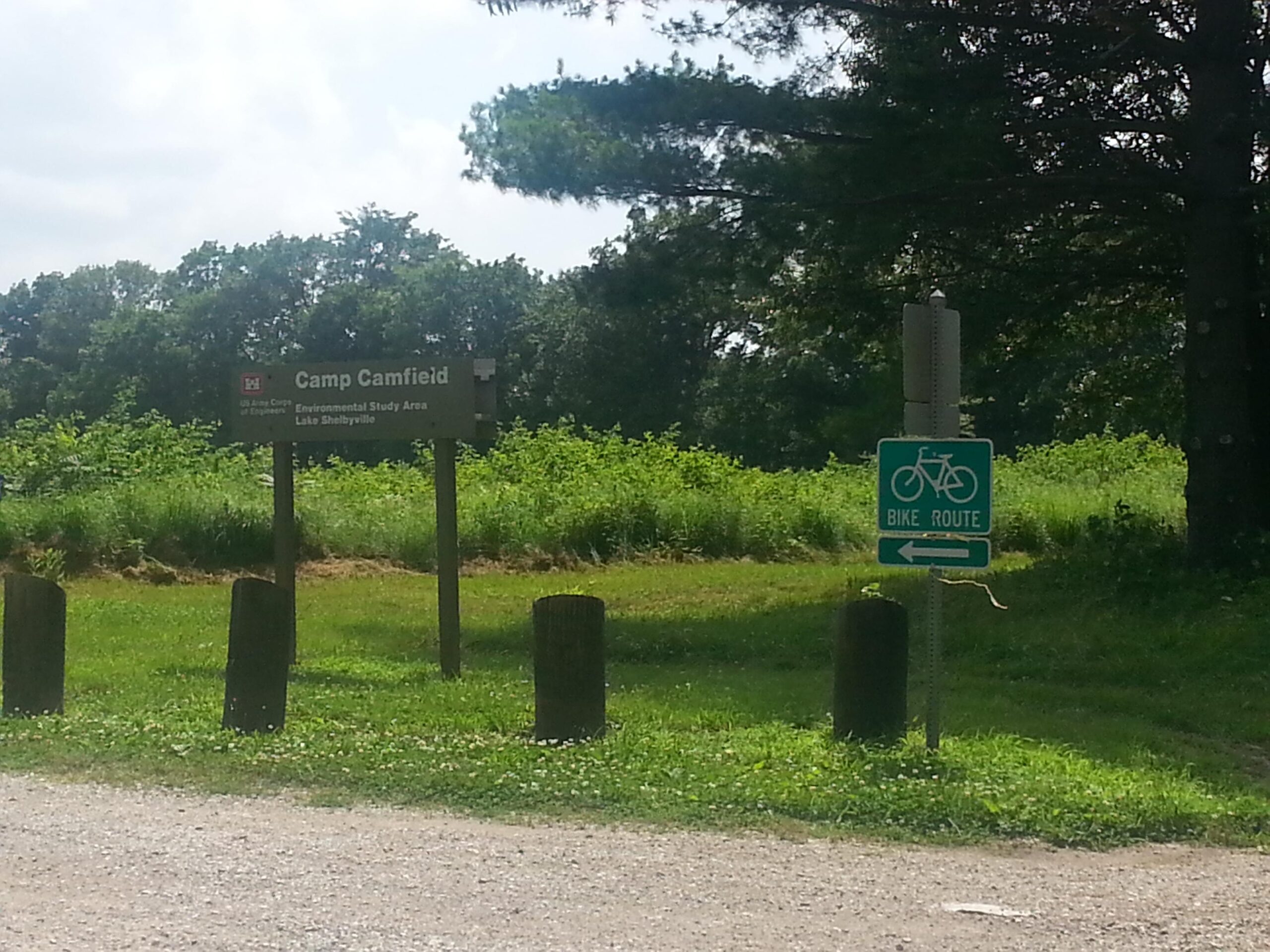 Sign for Camp Camfield, an Environmental Study Area at Lake Shelbyville, with a nearby bike route sign, surrounded by greenery and gravel path. Camp Camfield mountain bike trail.