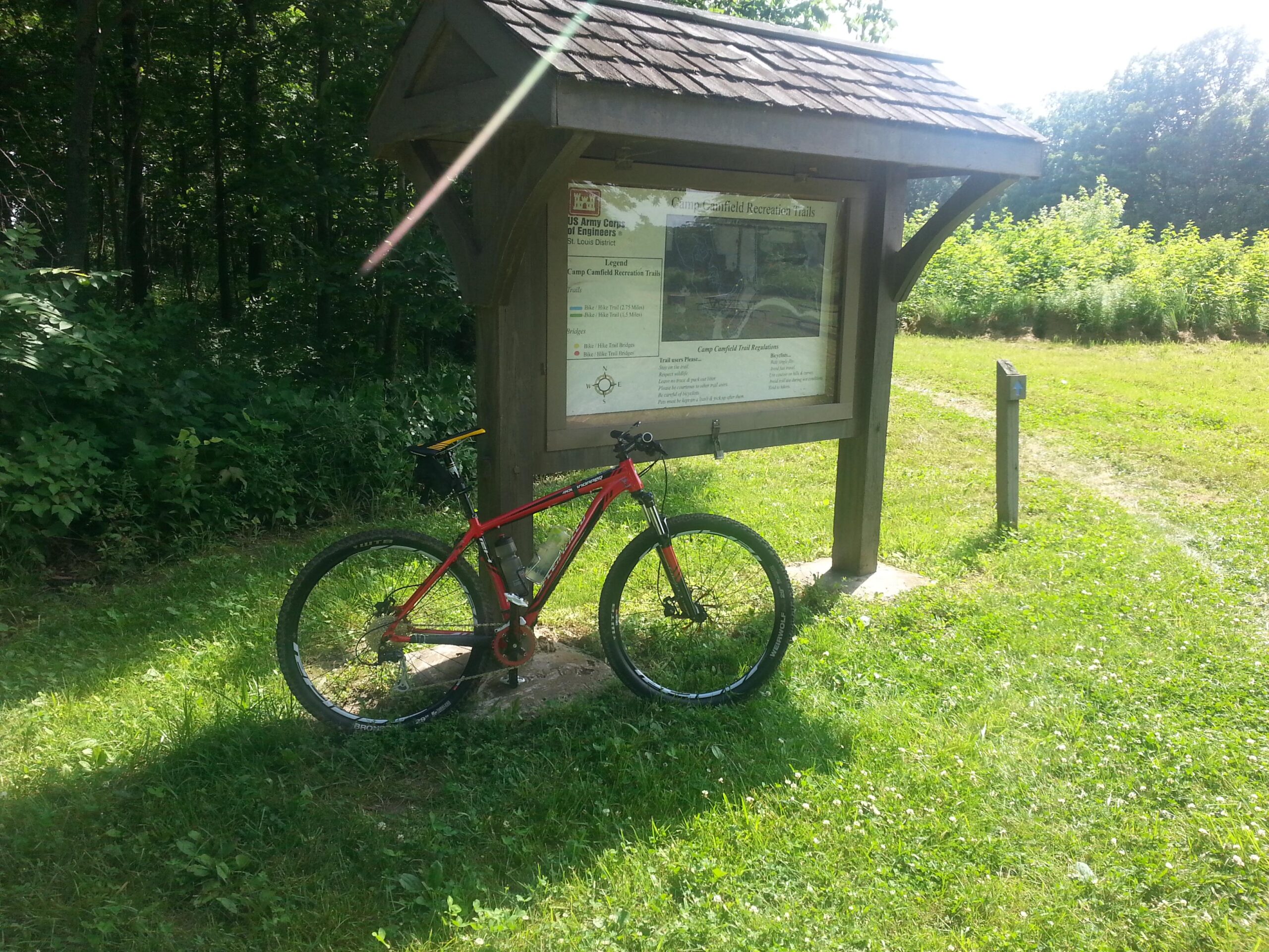 A red mountain bike is parked next to a wooden informational sign in a grassy area surrounded by trees. The sign provides details about the Camp Garfield Recreation Trails, and the scene is well-lit by sunlight on a clear day. Camp Camfield mountain bike trail.