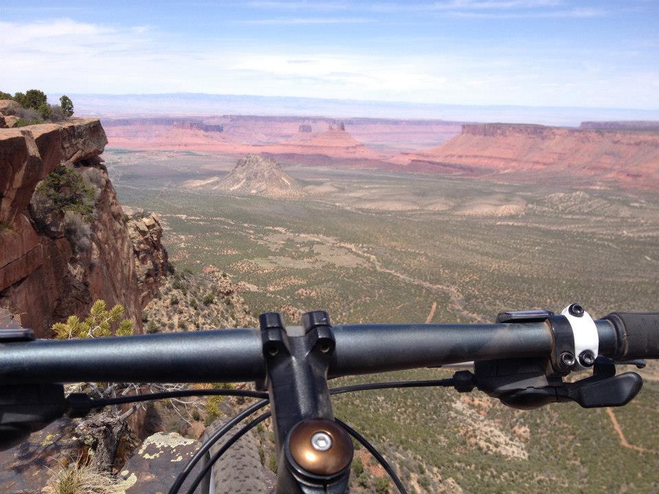 A mountain bike handlebar is in the foreground, with a breathtaking view of a desert landscape featuring cliffs, mesas, and a wide valley under a bright blue sky. The scene captures the natural beauty and rugged terrain of the region. Porcupine Rim mountain bike trail.