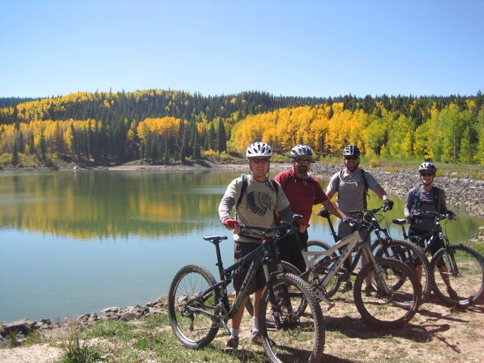 Four mountain bikers pose together by a serene lake, surrounded by vibrant autumn foliage. They are wearing helmets and biking gear, with bicycles in the foreground. The reflection of the colorful trees can be seen in the calm water. The sky is clear and blue, emphasizing the beauty of the outdoor setting. West Bench Trail mountain bike trail.