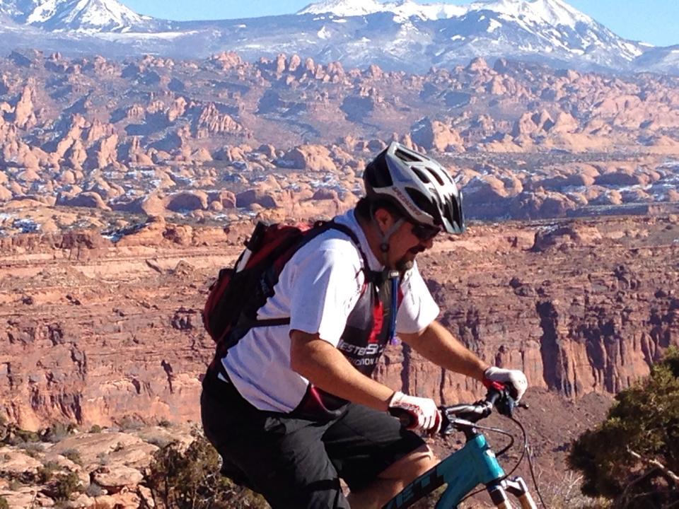 A mountain biker rides along a rugged trail with a dramatic rocky landscape and snow-capped mountains in the background. The cyclist is wearing a helmet and a cycling jersey, focused on navigating the terrain. The scene captures the essence of outdoor adventure and the beauty of nature. Amasa Back Trail mountain bike trail.