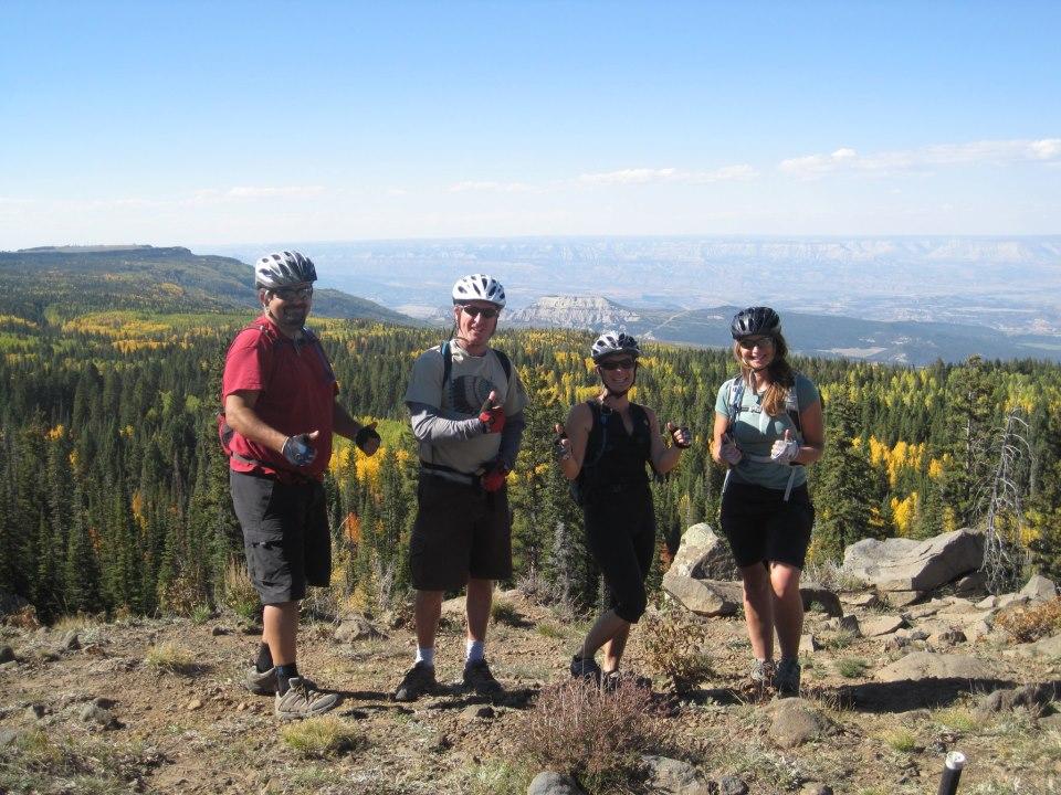 Four mountain bikers in helmets stand on a rocky outcrop with a panoramic view of a forested landscape and distant mountains. They are smiling and giving thumbs up, showcasing a sense of adventure and enjoyment in the outdoors. The trees display autumn colors, adding vibrancy to the scene. West Bench Trail mountain bike trail.