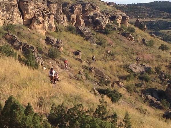 Hikers navigating a rocky terrain on a hillside, surrounded by grass and sparse vegetation, with cliffs in the background. Lunch Loops mountain bike trail.