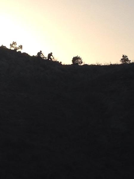 Silhouette of two cyclists atop a hill against a sunset sky, with trees in the background. Lunch Loops mountain bike trail.