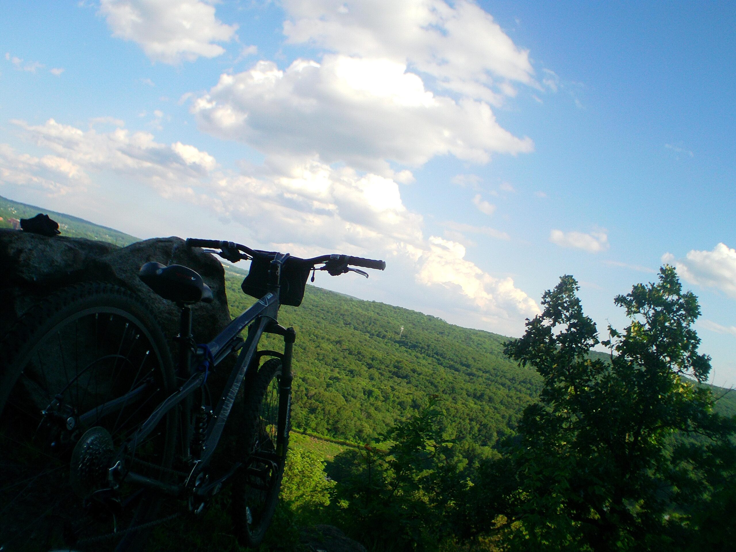 A mountain bike resting on a rock ledge, overlooking a lush green valley below, with a bright blue sky filled with fluffy white clouds. Allamuchy State Park-North mountain bike trail.