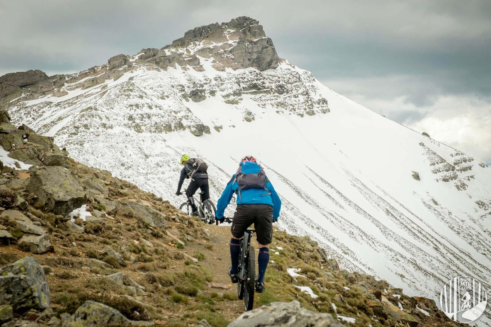 Two mountain bikers riding along a rocky trail in a mountainous area, with snow-covered peaks in the background and a cloudy sky overhead. The first rider, wearing a black helmet and gear, pedals ahead, while the second rider, dressed in a bright blue jacket, follows closely behind. The landscape is rugged, showcasing natural terrain and patches of grass. Pico Urbion mountain bike trail.