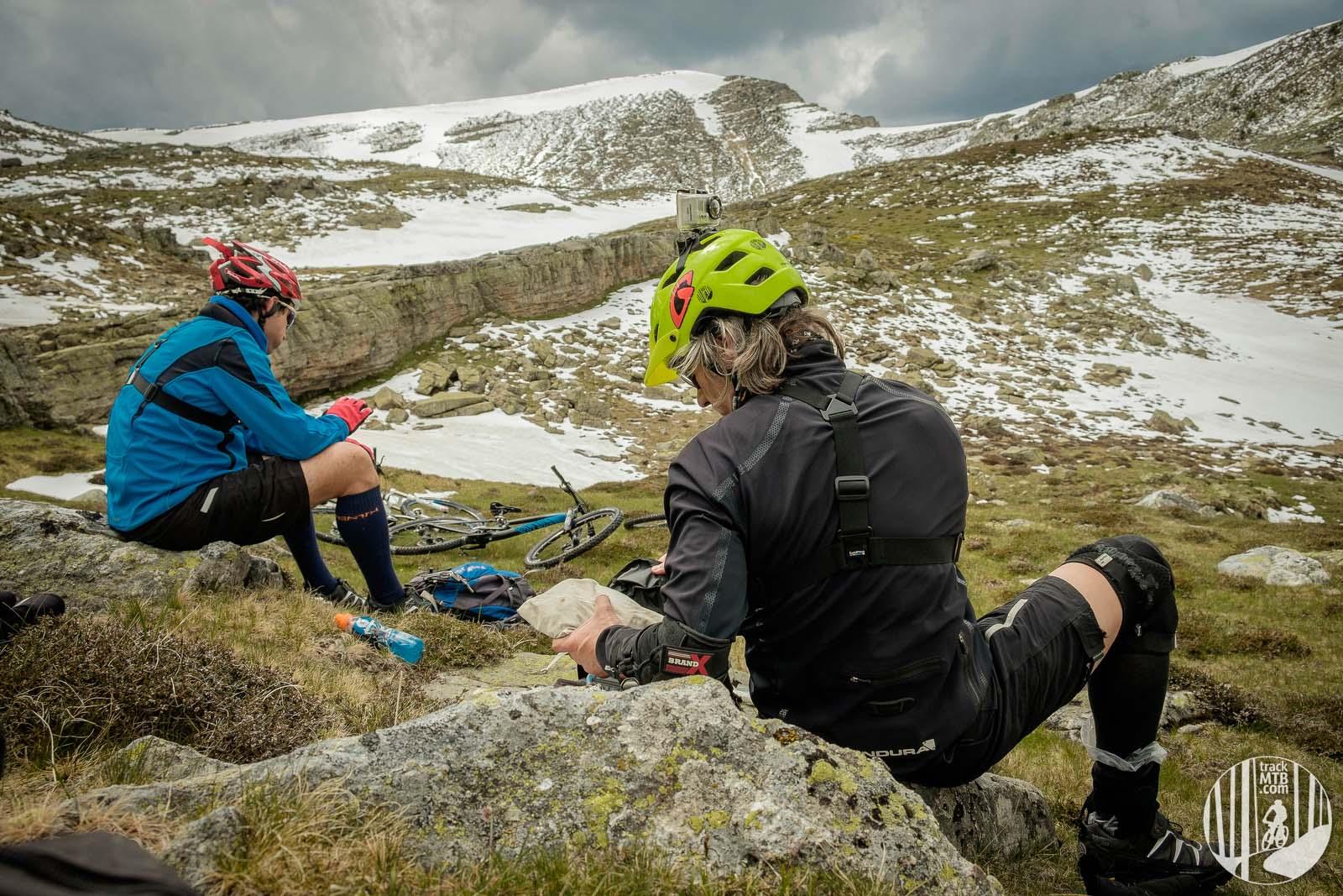 Two mountain bikers rest on rocky terrain in a mountainous area, surrounded by patches of snow and grass. One biker, wearing a blue jacket and red gloves, sits on a rock, using a mobile device. The other, dressed in black with a green helmet, is seated nearby, focused on a small object. Their bicycles are positioned on the ground beside them, while the cloudy sky looms above. Pico Urbion mountain bike trail.