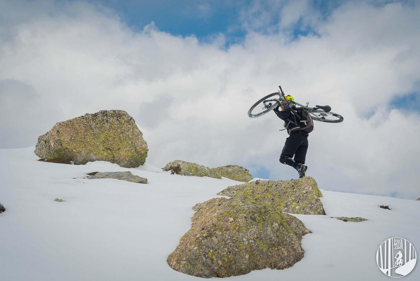 A biker carrying a mountain bike on their shoulder while traversing snow-covered rocks under a cloudy sky. Pico Urbion mountain bike trail.