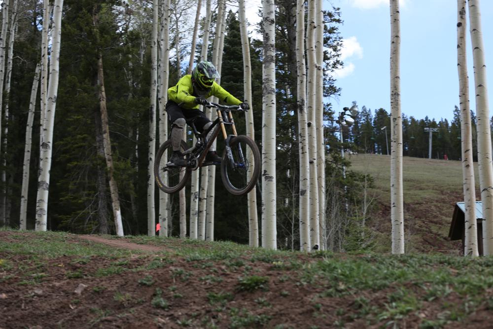 A cyclist in a bright yellow jacket performs a jump on a mountain bike, surrounded by tall white aspen trees. The scene captures the action of mountain biking on a dirt path with a grassy area in the foreground and a hilly landscape in the background. Angel Fire Bike Park mountain bike trail.