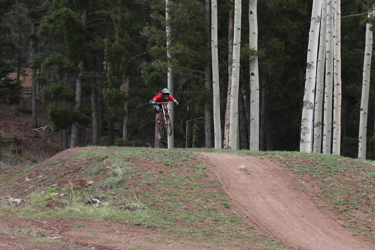 A mountain biker in a red jersey performs a jump on a dirt track surrounded by tall green trees. The bike is airborne as the rider prepares to land on the downhill slope. Angel Fire Bike Park mountain bike trail.