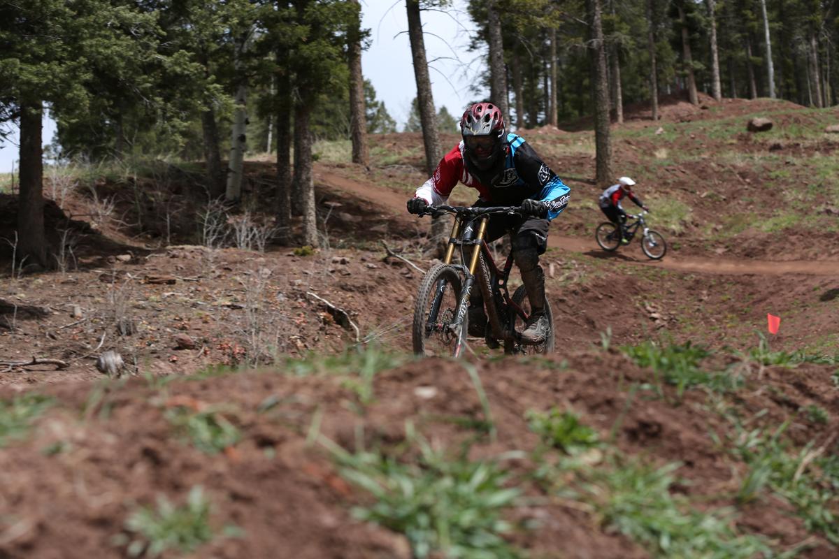 Mountain bikers riding on a dirt trail in a forested area. One rider is in the foreground, navigating a sloped section of the trail, while another rider is visible in the background on a different part of the path. The scene features trees and rocky terrain, with some grassy patches. Angel Fire Bike Park mountain bike trail.