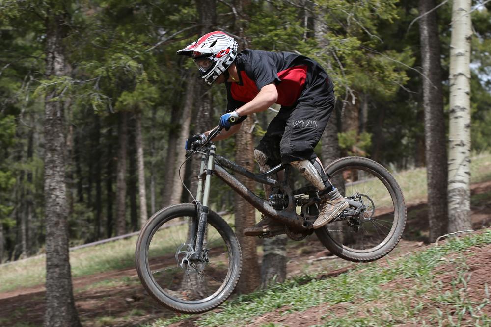 A person wearing a helmet, gloves, and protective gear jumps on a mountain bike while riding through a forested area. The bike is airborne, and the surrounding trees and greenery create a natural setting. Angel Fire Bike Park mountain bike trail.
