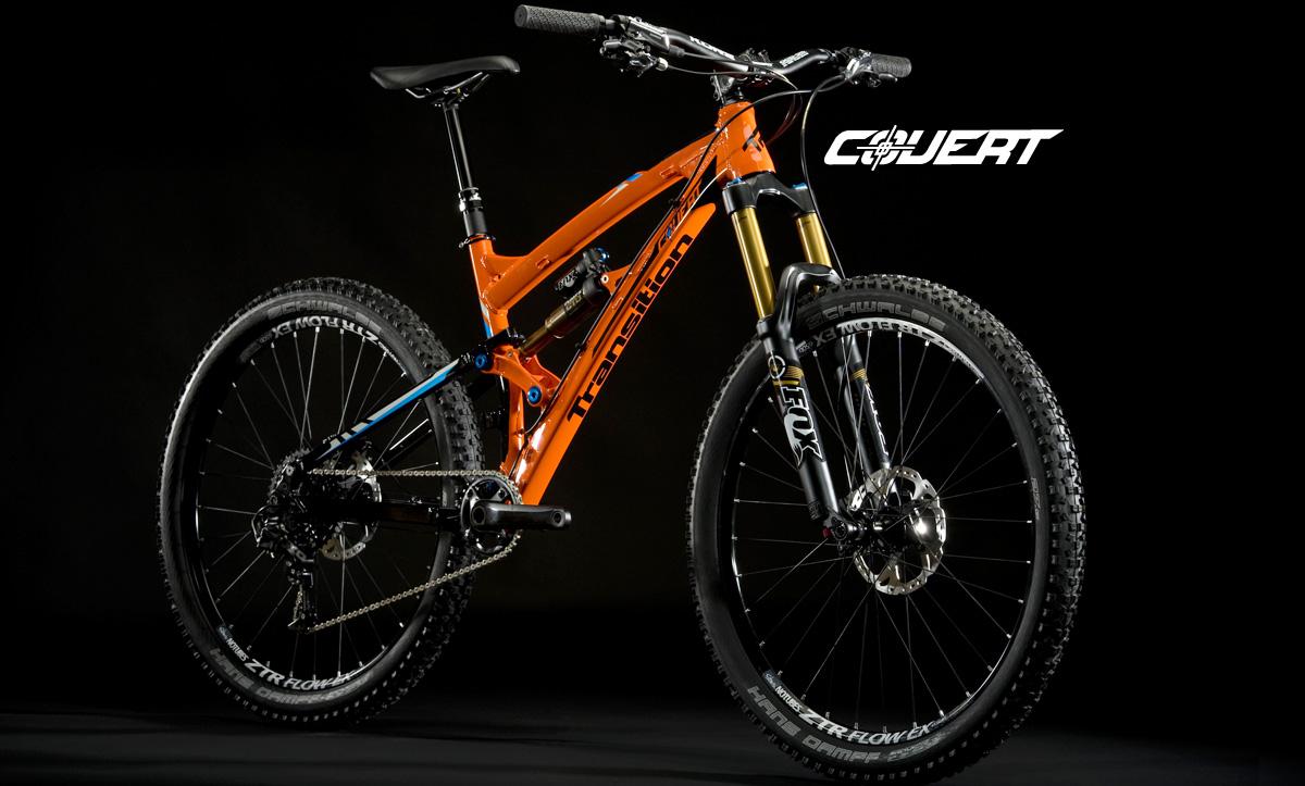Transition Covert 27.5: Mountain bike with an orange frame and blue accents, featuring a front suspension fork and thick tires, displayed against a black background.