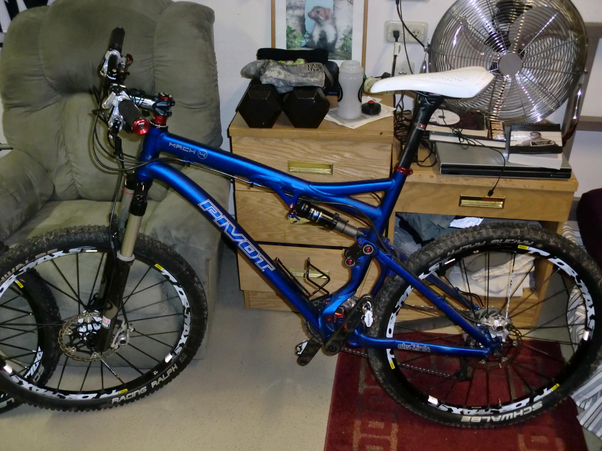 Pivot Mach 4: A blue mountain bike resting in a room with a green armchair in the background. The bike features large tires and a sleek frame, positioned next to a wooden chest of drawers and a fan. The setting suggests a casual indoor environment, possibly a personal living space or workshop.