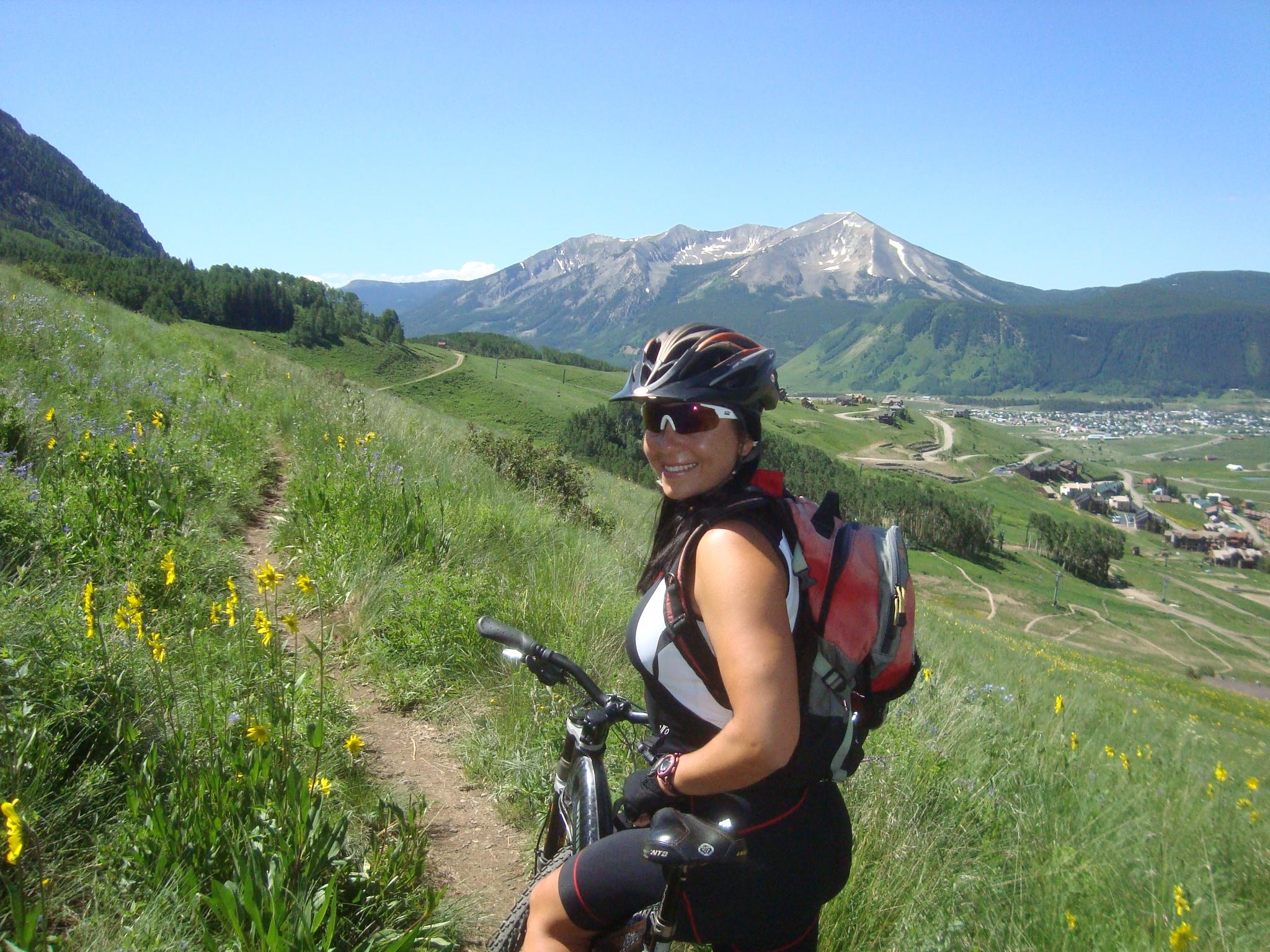 A woman wearing a helmet and sunglasses smiles while standing next to her mountain bike on a grassy trail. In the background, there are mountains and a clear blue sky, with a view of a village in the valley below. Wildflowers bloom along the path, creating a vibrant natural setting. Snodgrass mountain bike trail.