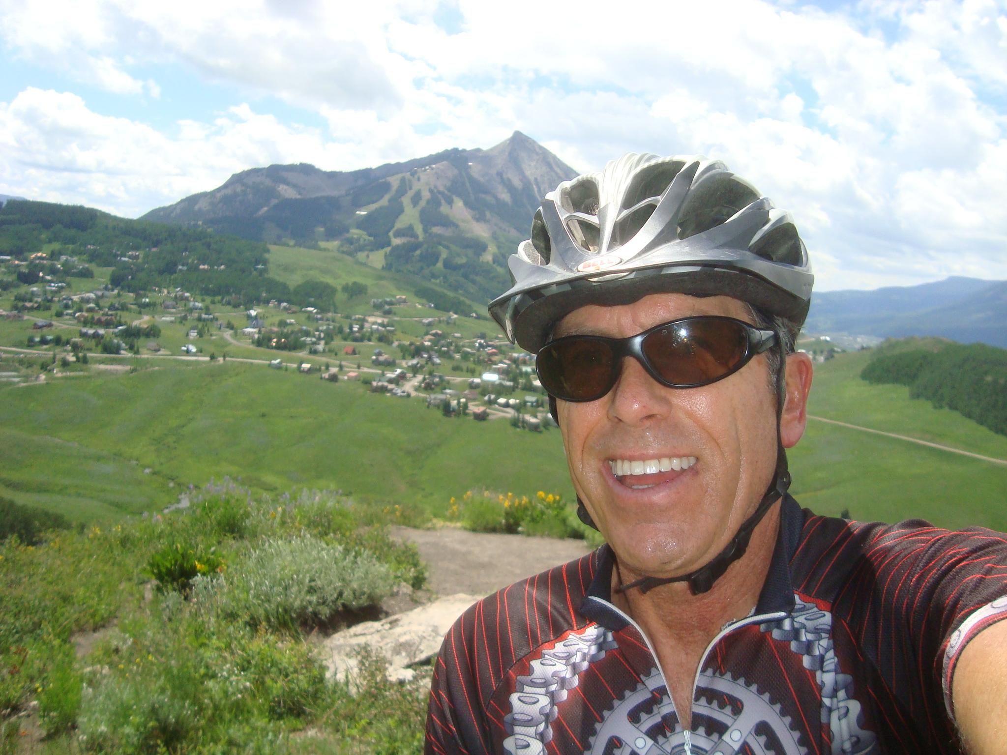 A smiling cyclist wearing a helmet and sunglasses takes a selfie against a backdrop of green hills, a small town, and a mountain range under a partly cloudy sky. Snodgrass mountain bike trail.