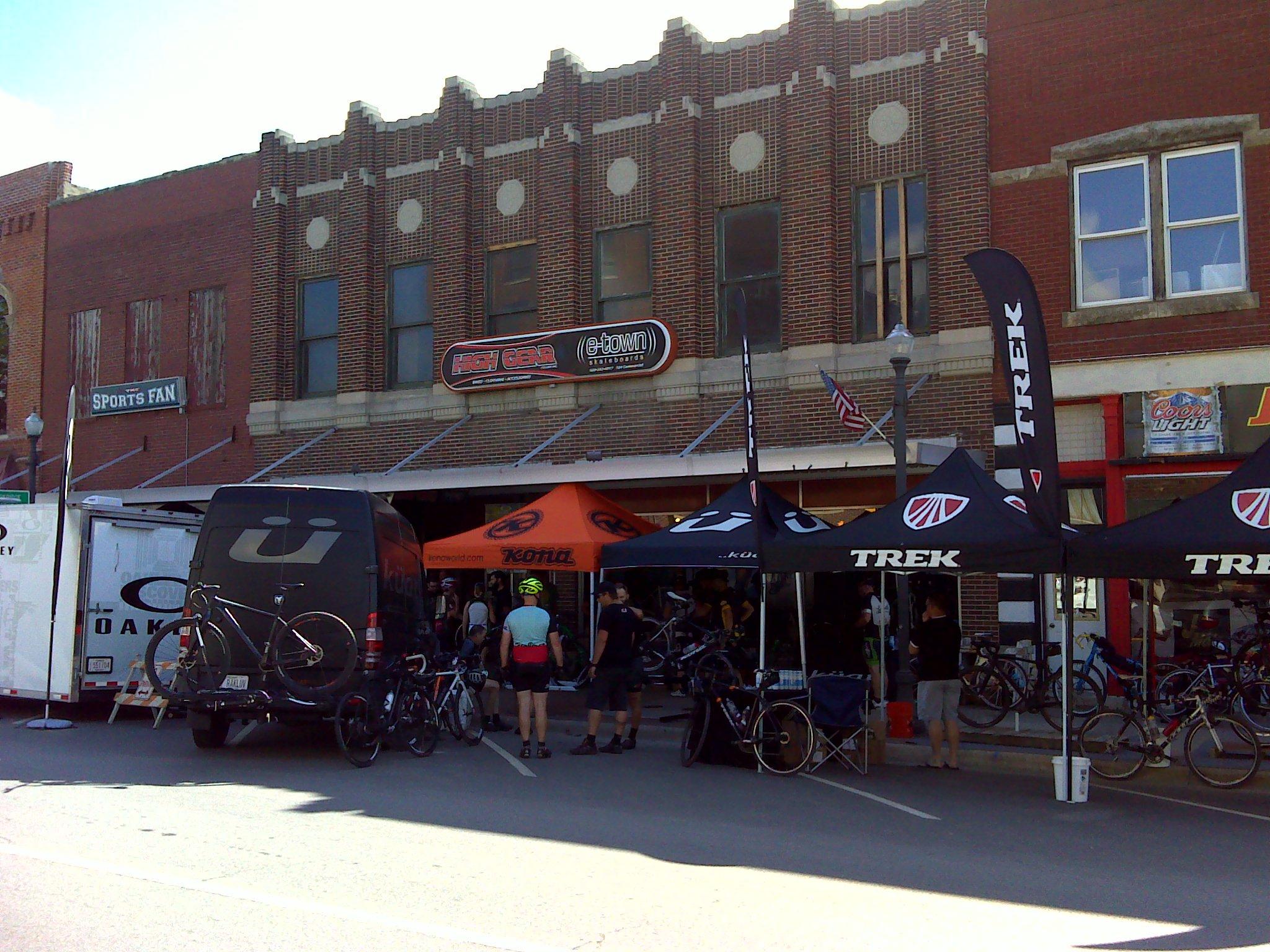 A bustling outdoor bike shop event featuring various tents and displays. The scene includes a van with bicycles attached to its rear, a tent displaying the Kona brand, and several Trek tents on display. People are gathered outside, engaging with bicycles and equipment. The backdrop features a historic brick building with storefronts and an American flag.