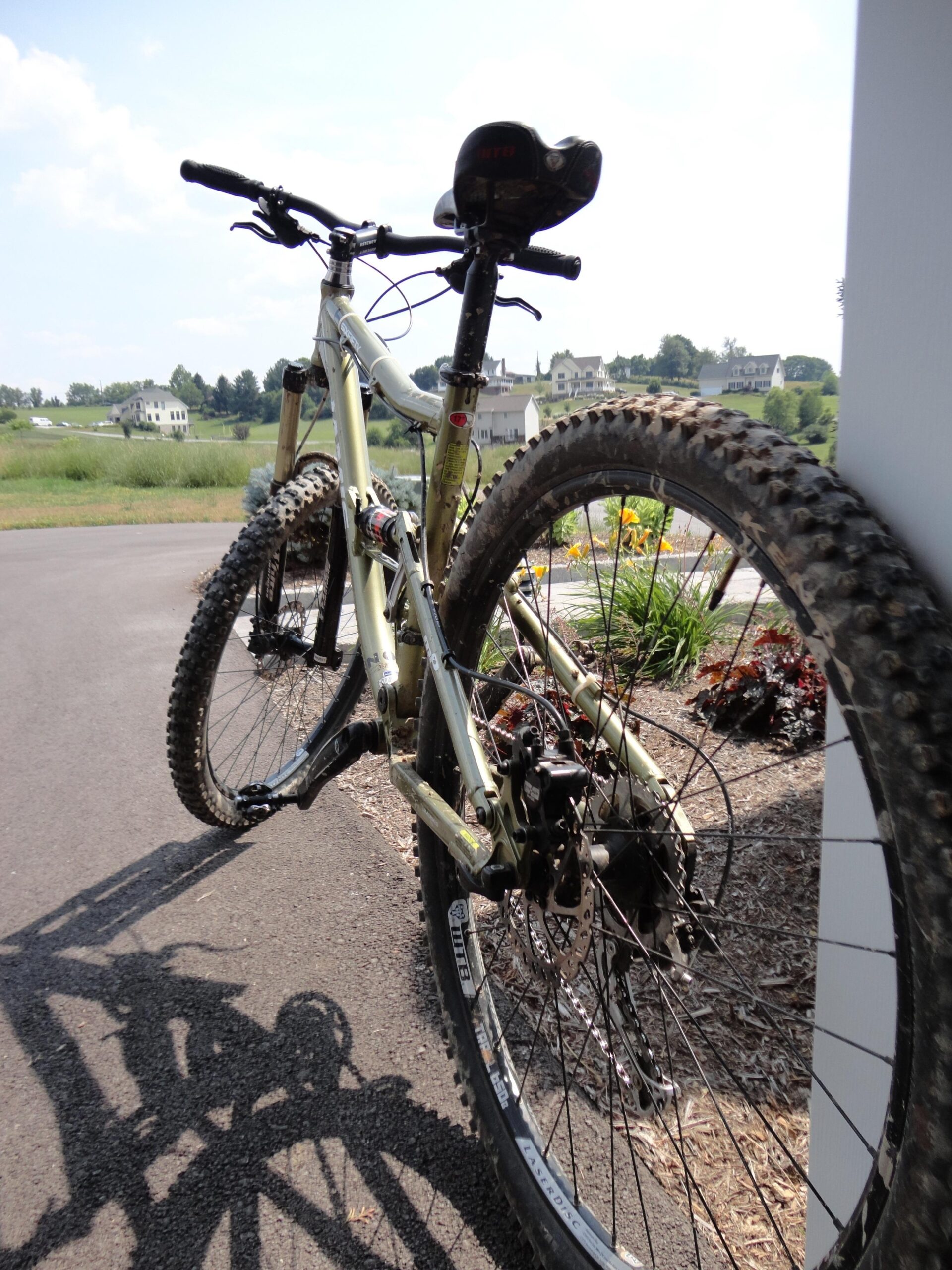 A close-up view of a mountain bike parked on a paved pathway, with the foreground highlighting the rear wheel and drivetrain. The background features a scenic landscape of green hills and houses under a clear sky. Pandapas Pond / Poverty Creek mountain bike trail.
