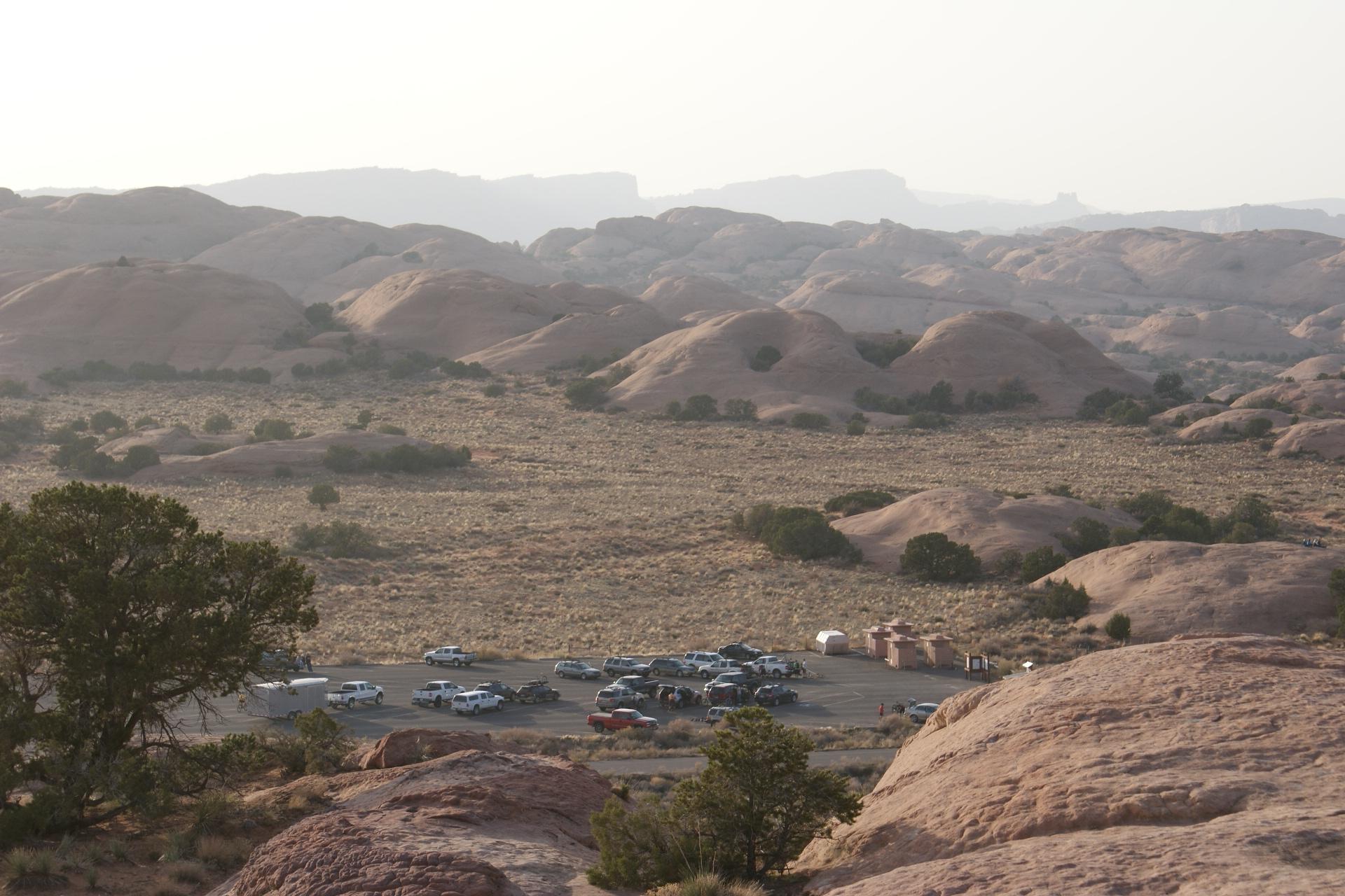 An expansive desert landscape featuring rounded rock formations and sparse vegetation. In the foreground, a small parking lot with several vehicles is visible, surrounded by low shrubs and rocky terrain. The background shows additional hills and distant mountains under a hazy sky, indicating a serene, natural setting. Slickrock mountain bike trail.
