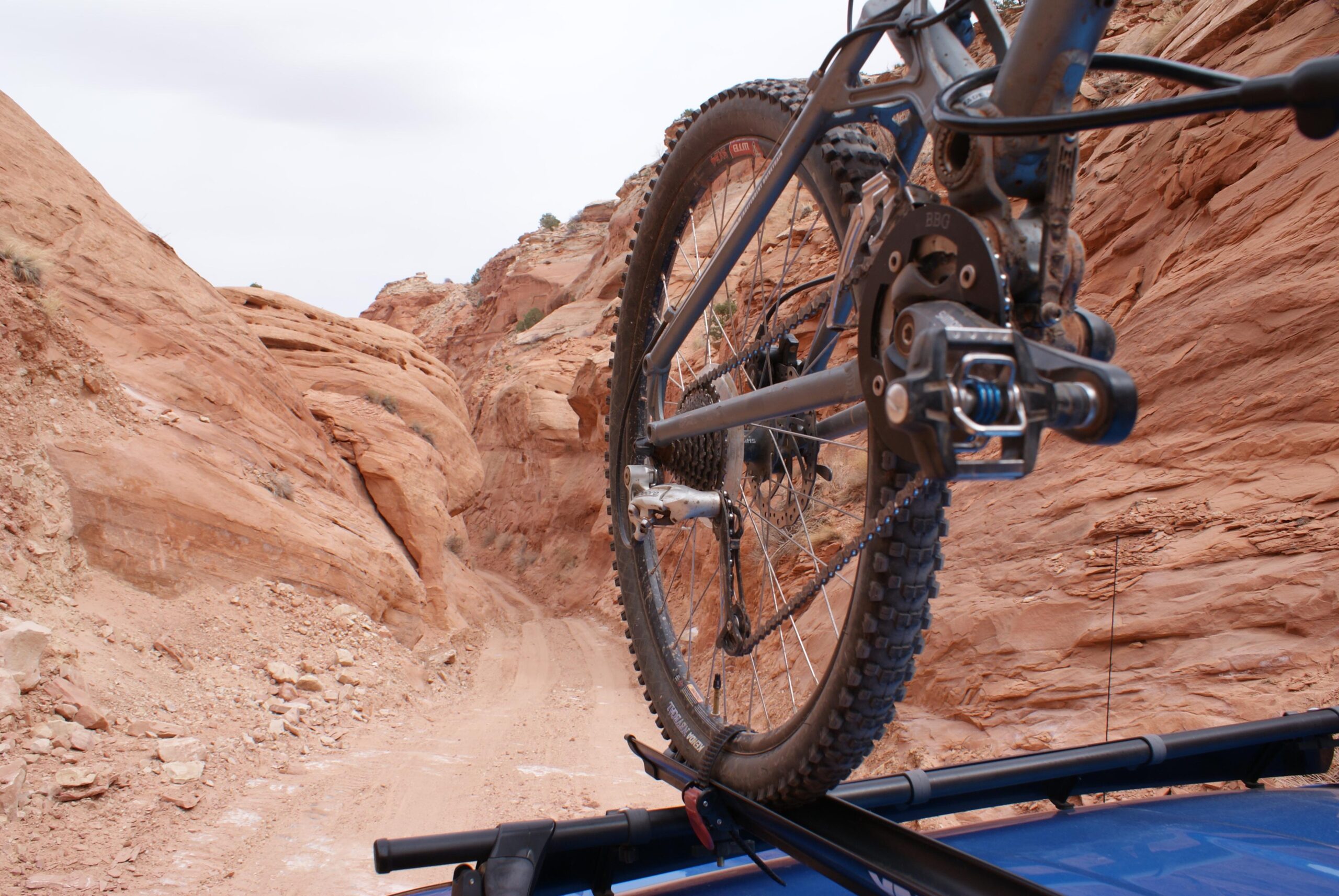 A close-up view of a mountain bike mounted on a rooftop rack, with a rugged dirt path and red rock formations in the background. The scene captures the outdoor adventure spirit, showcasing the bike's tire and gear mechanics as it overlooks a winding trail through the rocky landscape. Long Canyon 4x4 Road mountain bike trail.