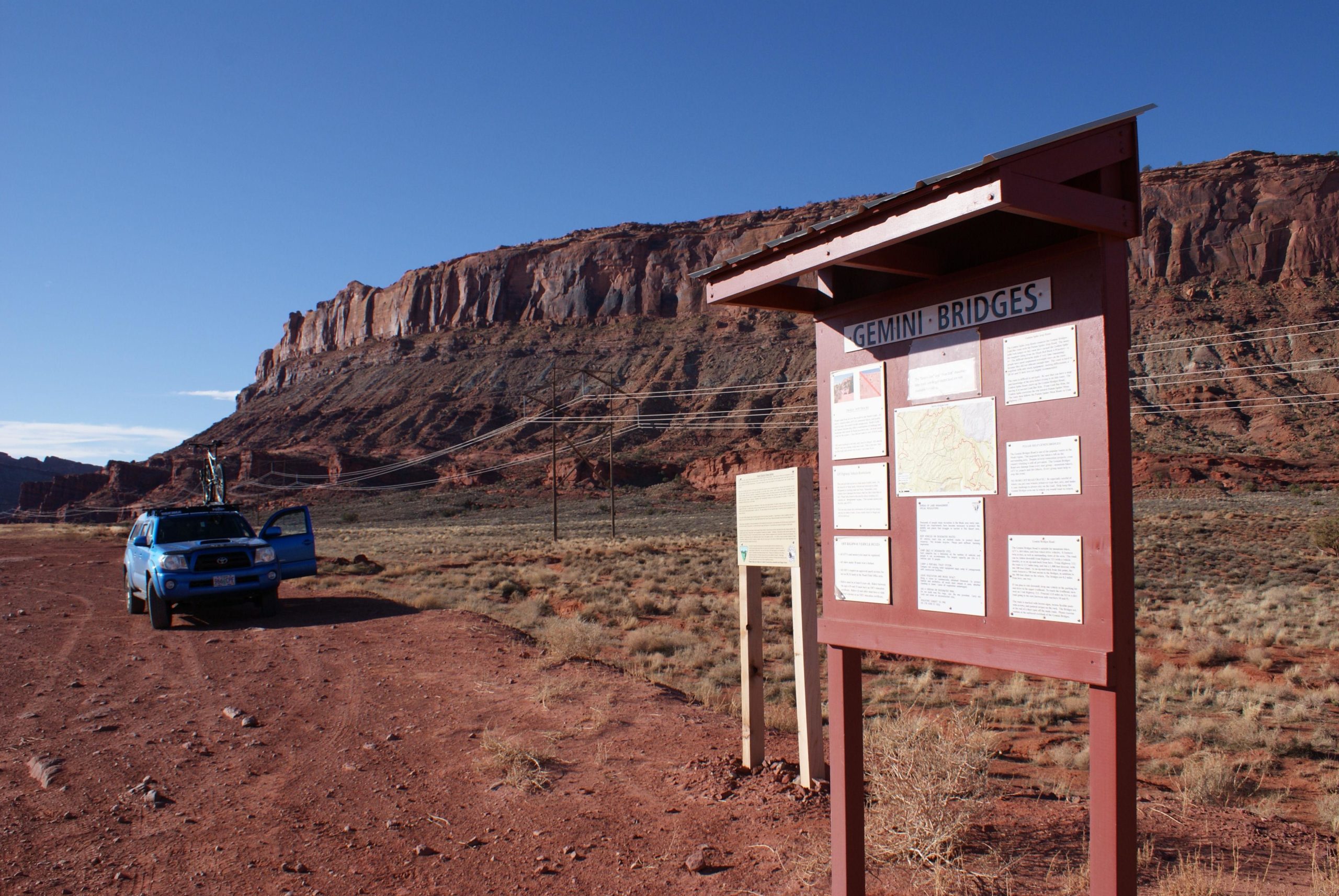 A blue SUV parked on a dirt road near a sign for Gemini Bridges, with informational panels about the area. Red rock cliffs rise in the background under a clear blue sky. Gemini Bridges mountain bike trail.