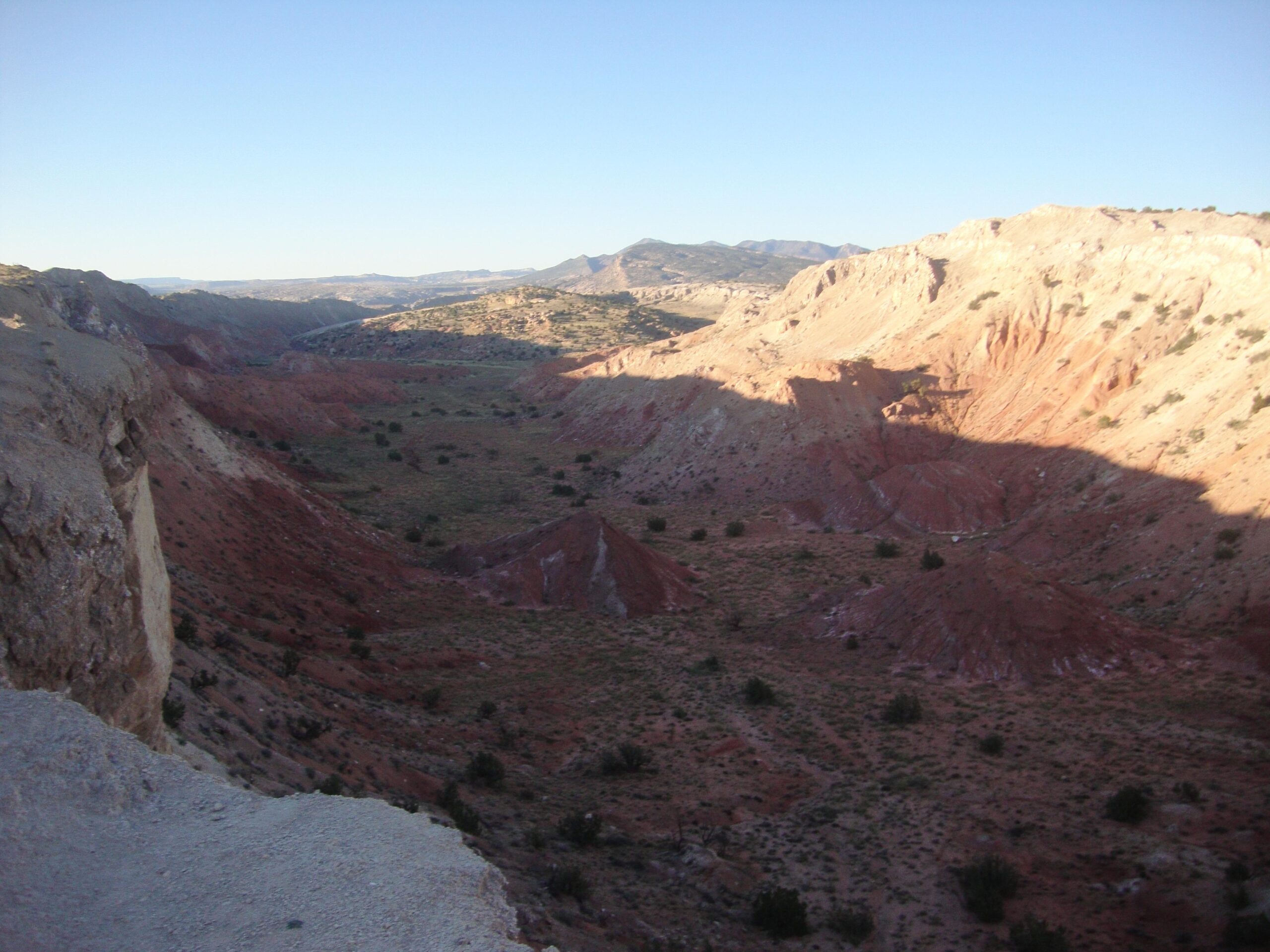 A panoramic view of a rocky landscape featuring red and brown hills and valleys under a clear blue sky. The terrain displays a mix of vegetation and rocky formations, with shadows highlighting the contours of the land. White Ridge Bike Trails mountain bike trail.