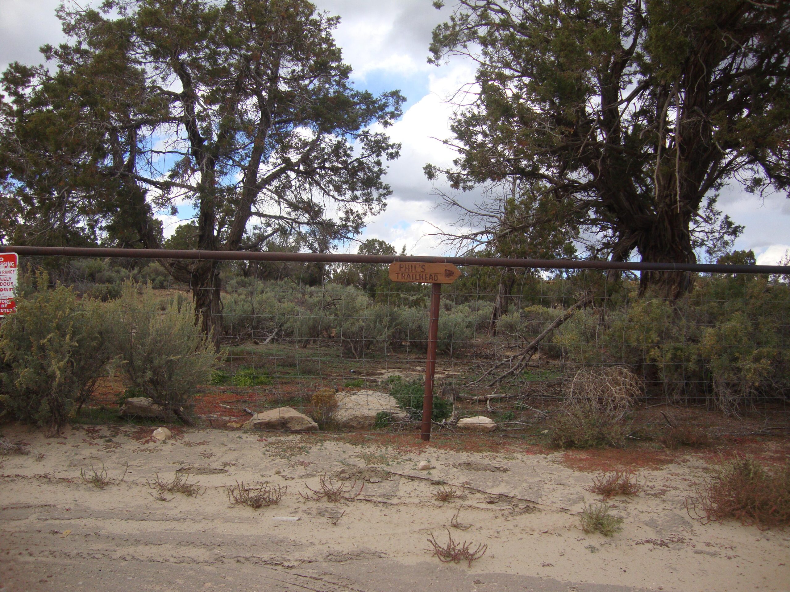 A dirt road leads up to a wooden sign for "Phil's Trailhead," surrounded by shrubs and trees. In the background, a fence is visible with a warning sign. The sky is partly cloudy, and the landscape features a mix of dry, sandy ground and green vegetation. Phil's World mountain bike trail.