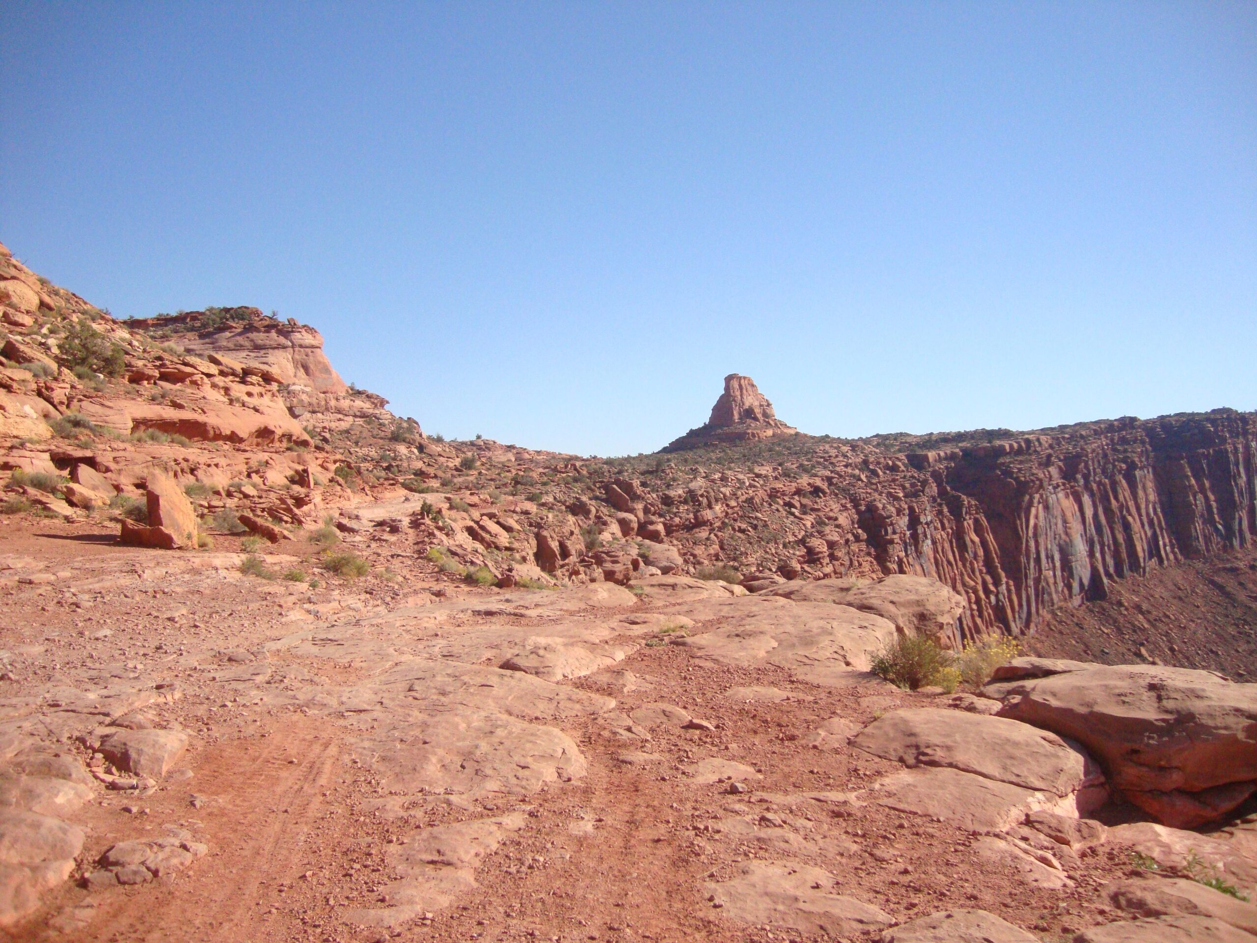 A rugged, dirt path winds through a landscape of red rock formations under a clear blue sky. In the distance, a prominent rock structure rises above the surrounding terrain, showcasing the natural beauty and geological features of a desert environment. Scrubby vegetation is sparsely scattered among the rocks, adding a touch of greenery to the arid scene. Amasa Back Trail mountain bike trail.