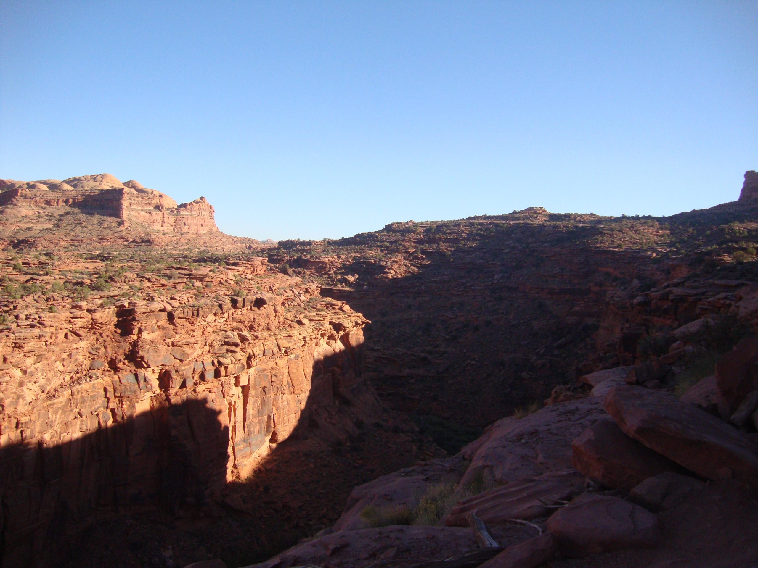 A scenic view of a rocky canyon under a clear blue sky, featuring steep cliff edges and rugged terrain. Shadows cast by the cliffs add depth to the landscape, while patches of green vegetation are visible among the rocks. Amasa Back Trail mountain bike trail.