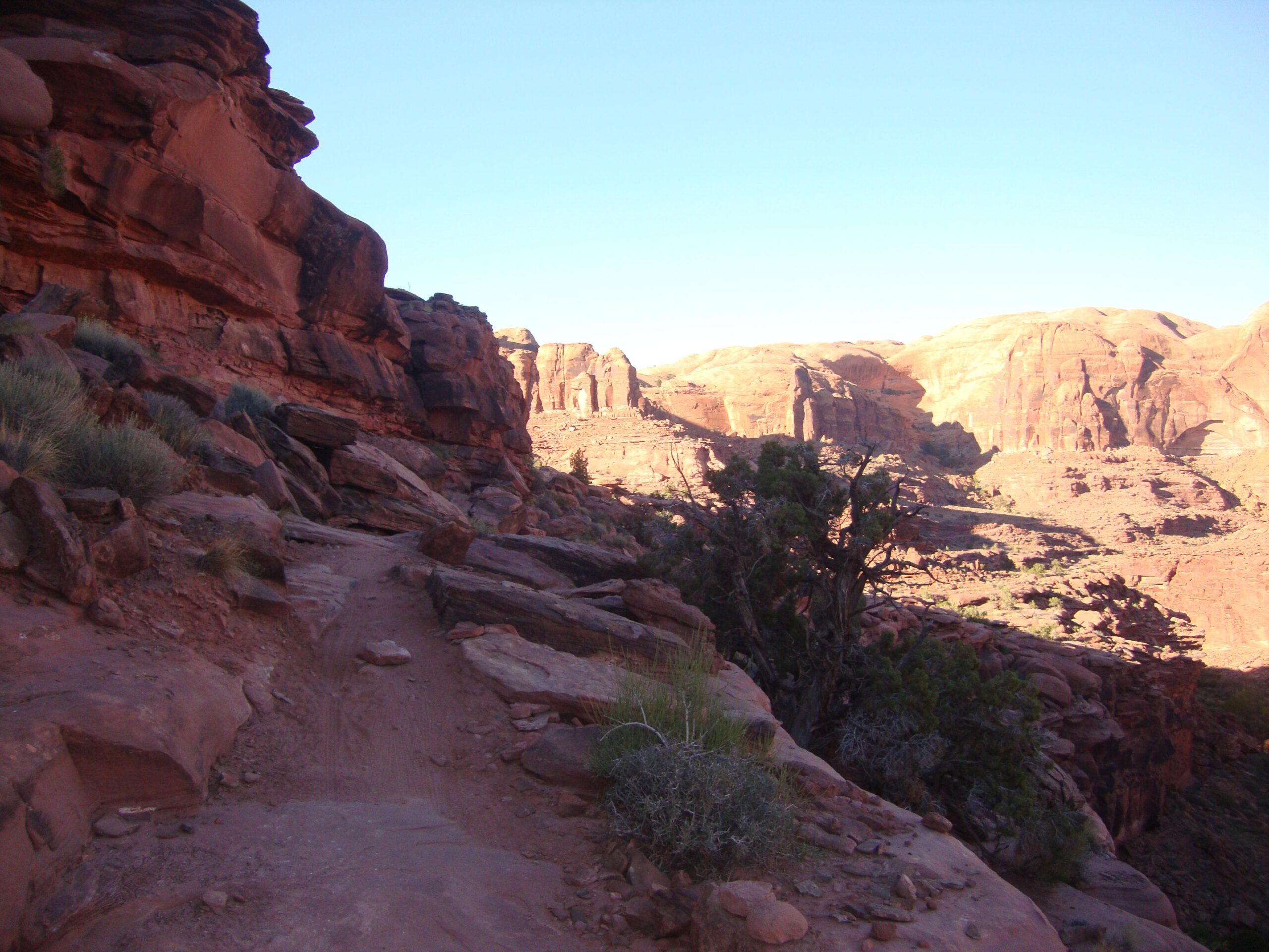 A narrow dirt path winding through red rock formations, with steep cliffs and greenery visible on the sides. The landscape is bathed in warm sunlight, highlighting the textures of the rocks and the shadows cast by the terrain. Amasa Back Trail mountain bike trail.
