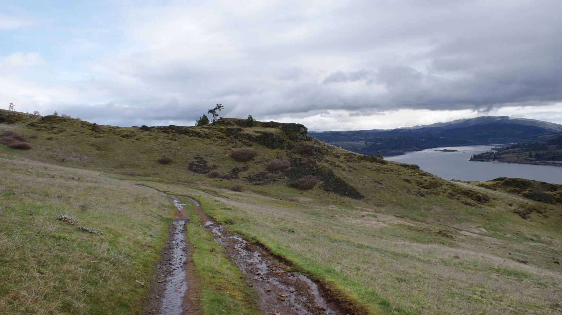 A winding dirt path leads through grassy hills, with a view of a river and distant mountains under a cloudy sky. Sparse vegetation, including shrubs and a few trees, dot the landscape. The scene captures a tranquil, natural environment. Syncline mountain bike trail.