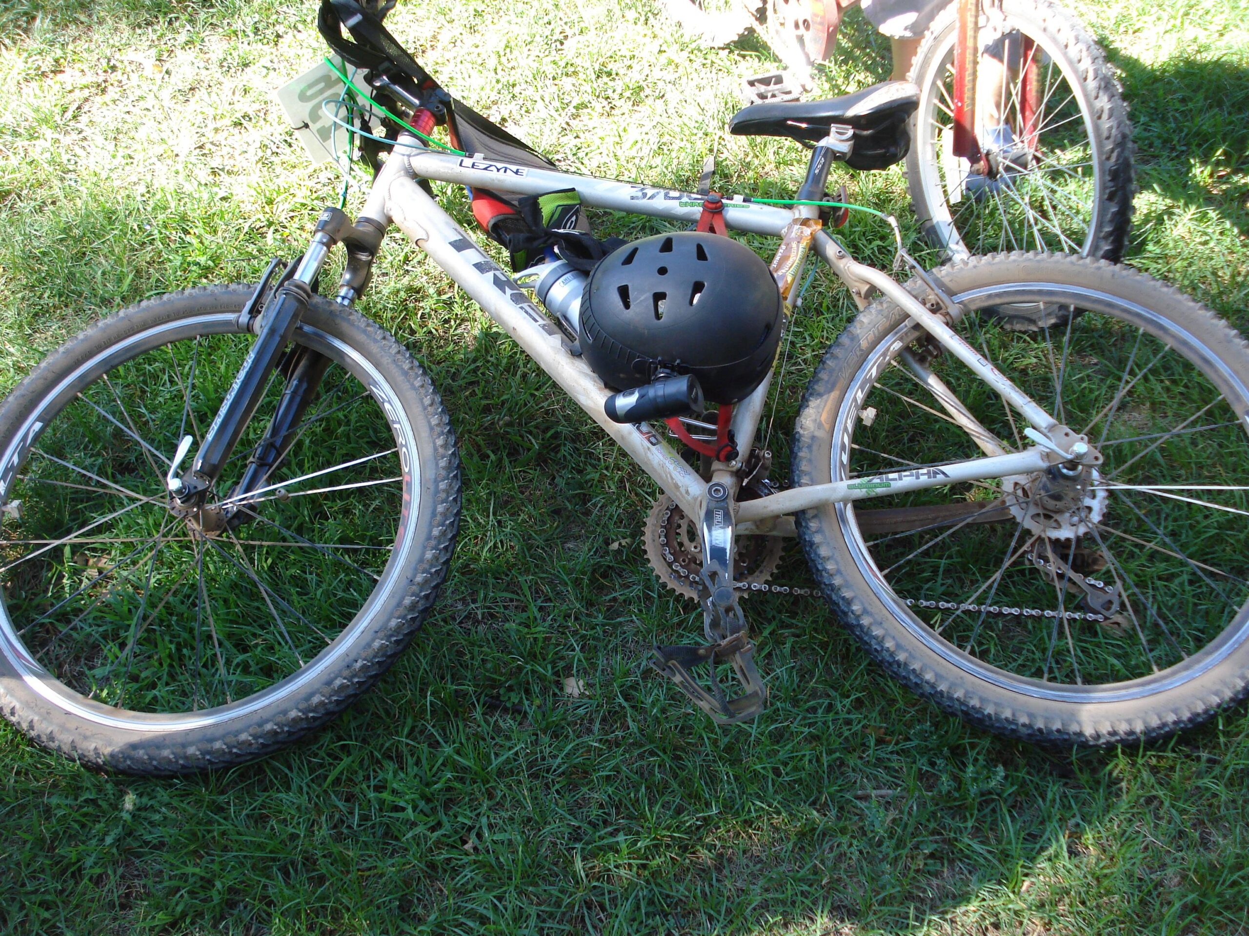 Trek 3700: A close-up image of a mountain bike resting on grass. The bike features a light-colored frame with visible dirt on the tires and components, and it's equipped with a helmet mounted on the frame. The bike's front wheel is angled to the side, showcasing its suspension system, while a second bike is partially visible in the background.