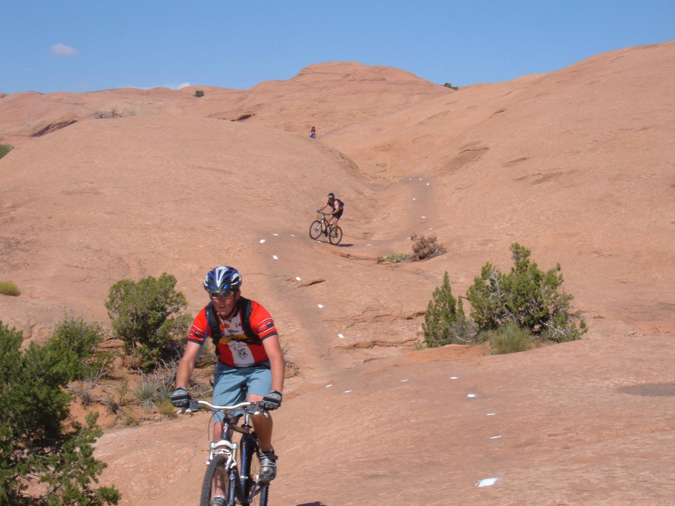 Two mountain bikers navigating a rocky terrain under a clear blue sky. The foreground features a biker wearing a red jersey and helmet, while another biker can be seen in the background. The landscape is characterized by smooth, sandstone formations and sparse vegetation. Slickrock mountain bike trail.