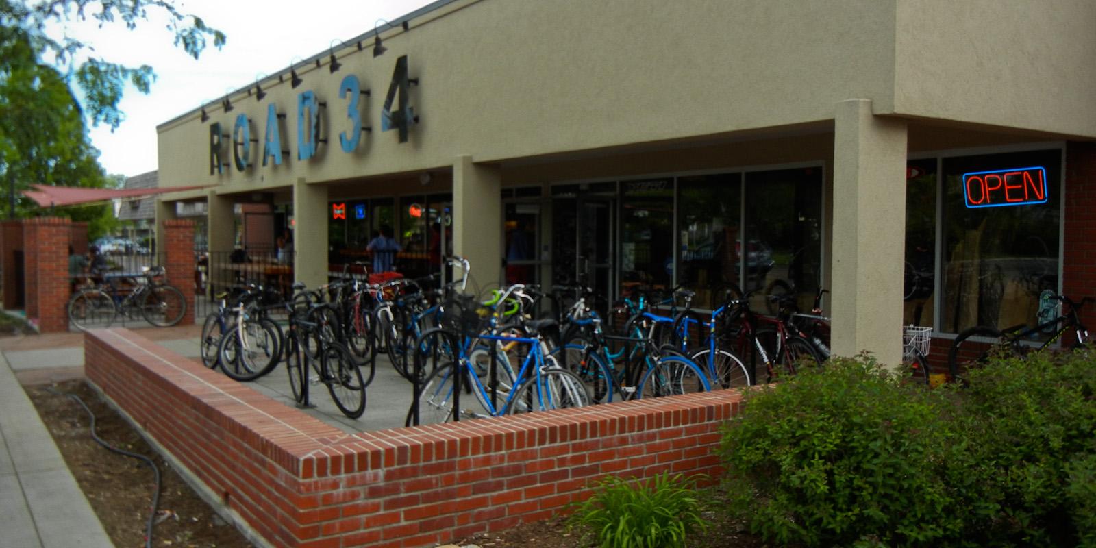 Alt text: Exterior view of a building with the sign "ROAD 34," featuring a variety of bicycles parked outside. The entrance has an illuminated "OPEN" sign, and there are some plants and a brick wall in the foreground.