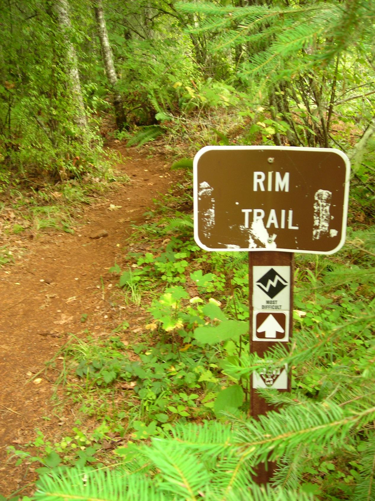 A wooden sign indicating the "Rim Trail," surrounded by lush green foliage. The sign features additional symbols indicating the trail's difficulty level and directional guidance, leading into a wooded area with a dirt path. Molalla River Recreation Corridor mountain bike trail.
