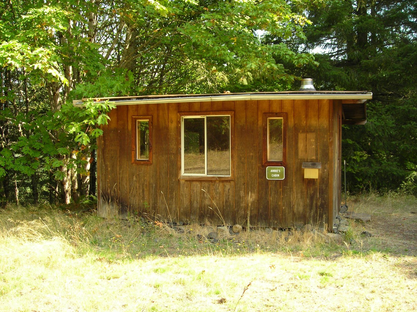 A small wooden cabin with a sign reading "Annie's Cabin" set in a grassy area, surrounded by trees. The cabin features two small windows and a larger front window. Sunlight filters through the foliage, creating a serene, rustic atmosphere. Molalla River Recreation Corridor mountain bike trail.