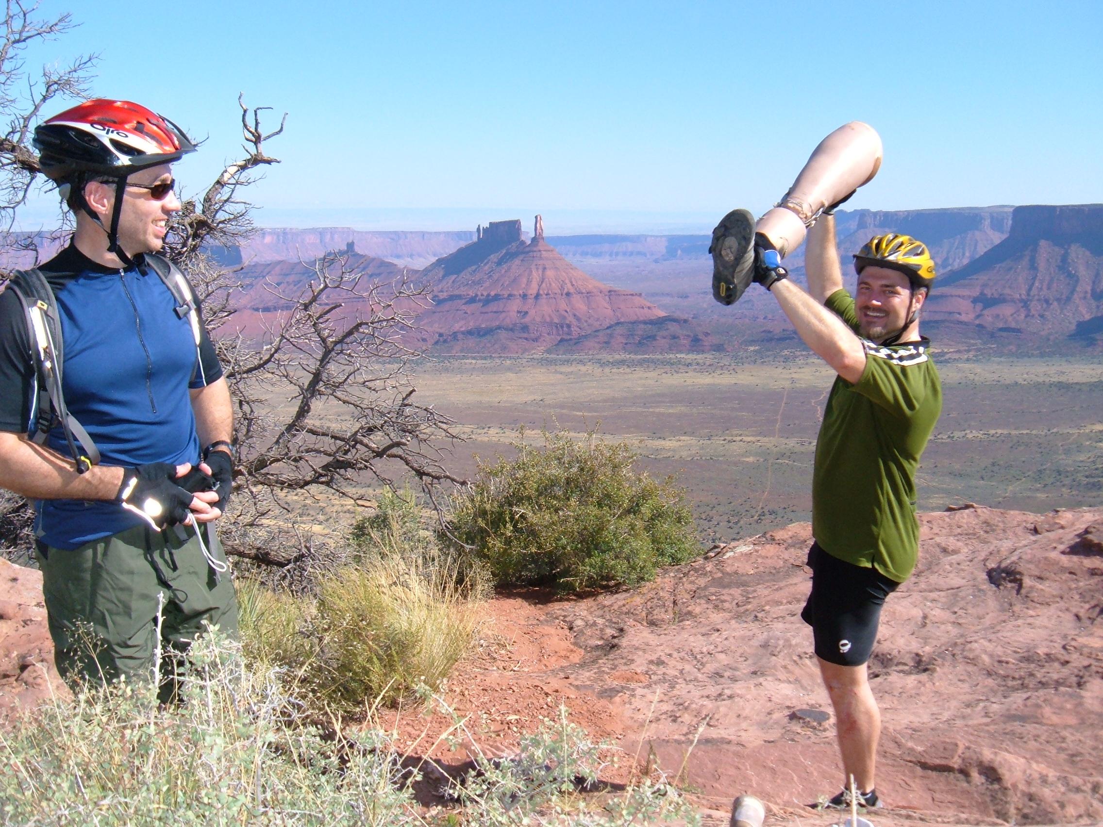 Two men are standing on a rocky terrain with a vast desert landscape in the background. One man, wearing a blue shirt and helmet, is looking at the other man, who is raising a prosthetic leg triumphantly above his head. They are both dressed in active wear, and the scene captures a moment of camaraderie and adventure against a backdrop of cliffs and mountains. Porcupine Rim mountain bike trail.