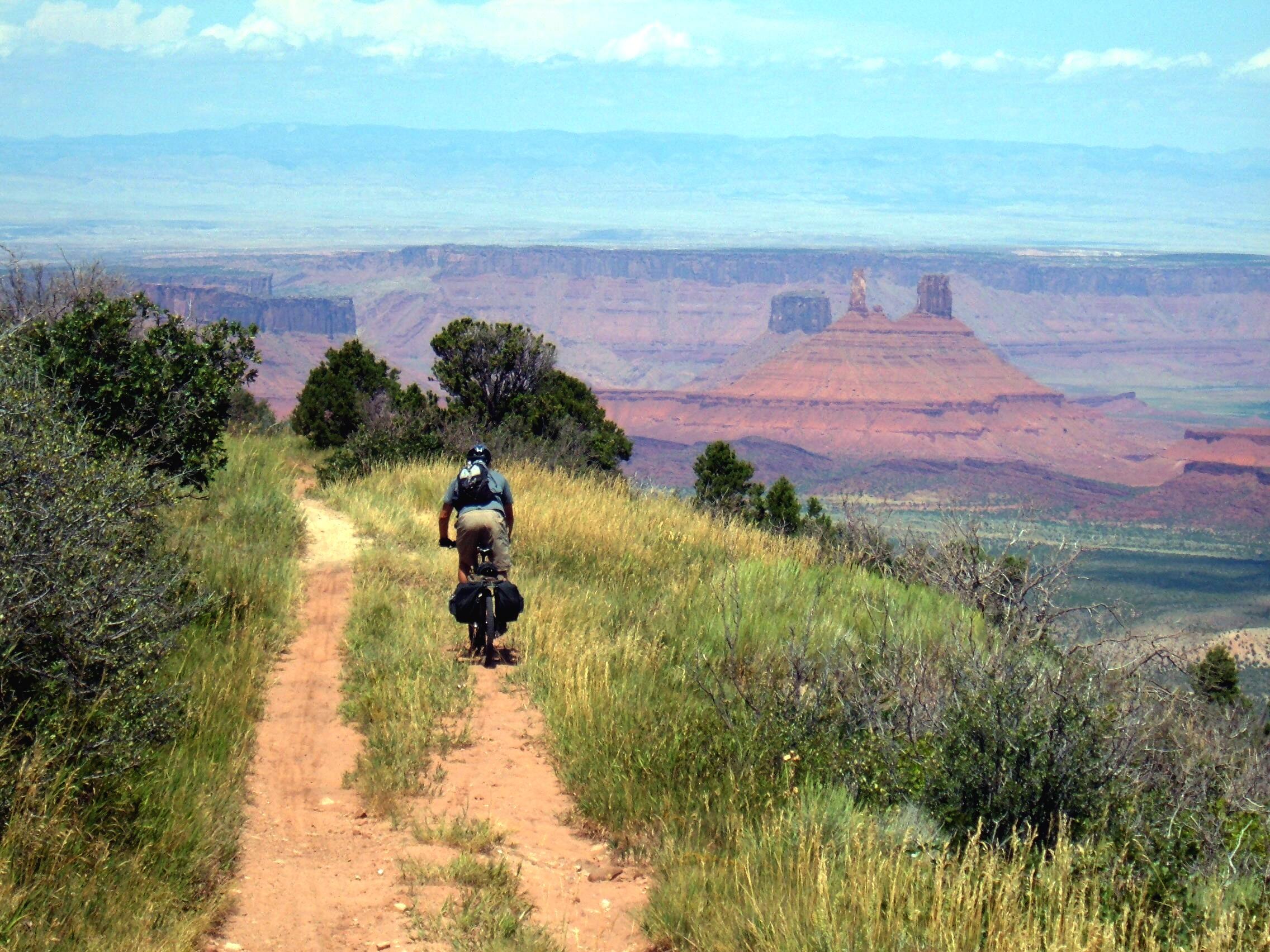 A person riding a bicycle along a dirt path surrounded by grass and shrubs, with a stunning view of red rock formations and distant mountains in the background under a partly cloudy sky. Kokopelli Trail mountain bike trail.