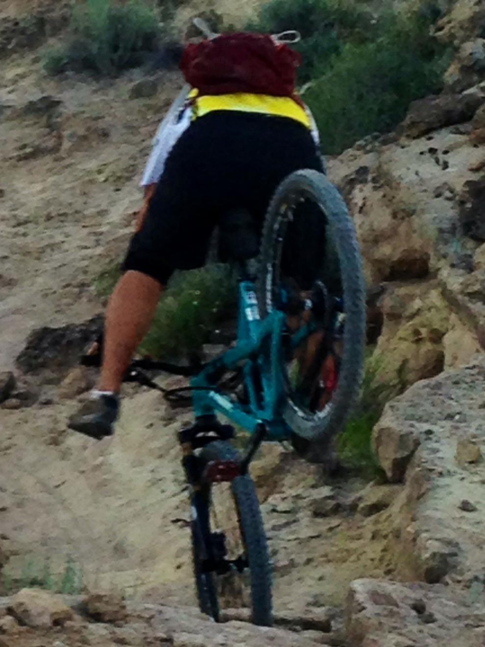 A mountain biker navigating a rocky terrain, leaning forward on their bike with one wheel elevated off the ground, showcasing an action-packed moment of trail riding. They are wearing a yellow and black cycling outfit and a red backpack. The background features a natural landscape with sparse vegetation and rocky surfaces. Lunch Loops mountain bike trail.