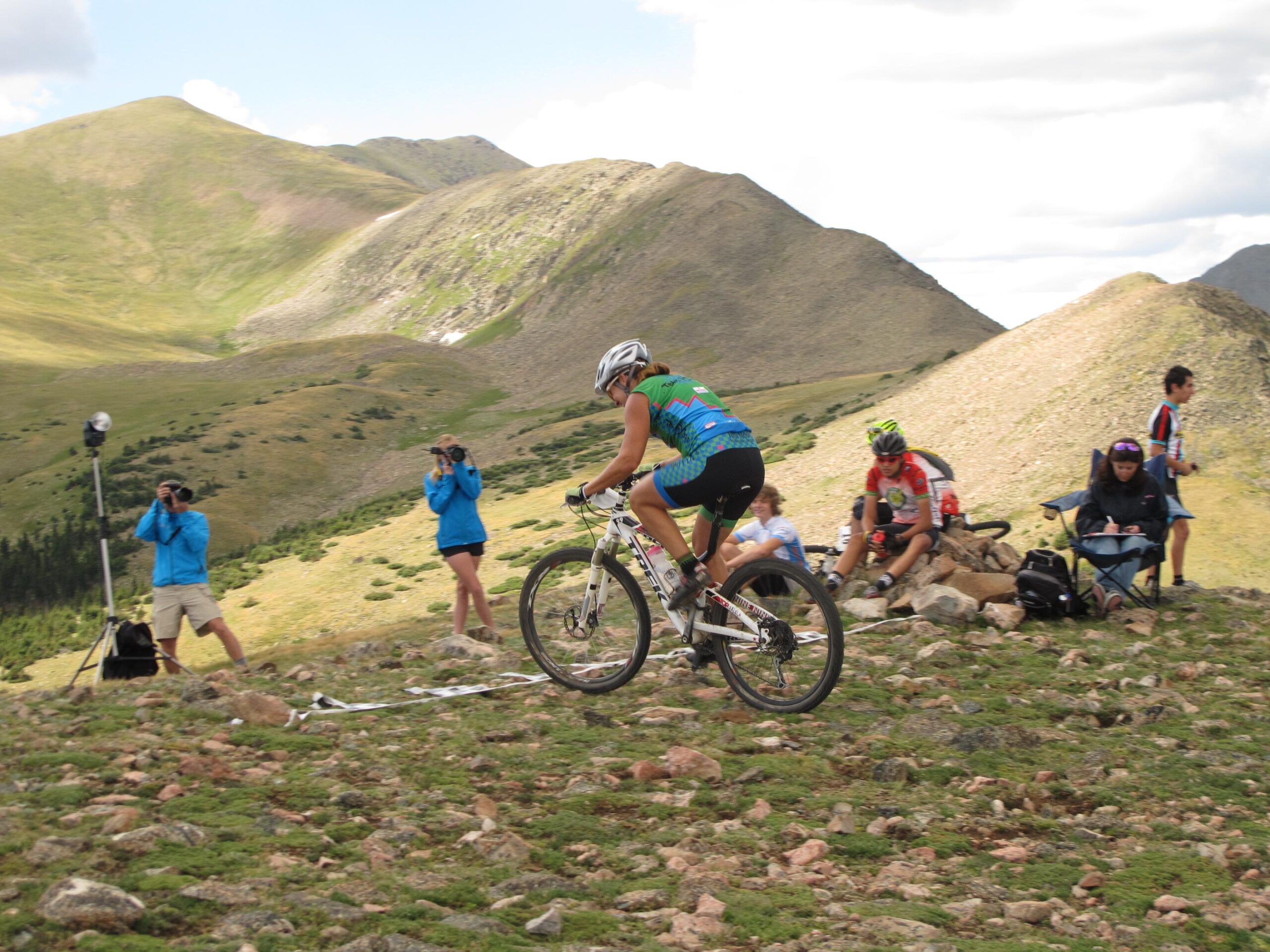 A mountain biker in a colorful jersey rides on a gravel path surrounded by rolling hills and mountains. Several spectators in blue jackets take photos, while others sit nearby, enjoying the view. The landscape features lush greenery and rocky terrain under a partly cloudy sky. Northside At Taos Ski Valley mountain bike trail.