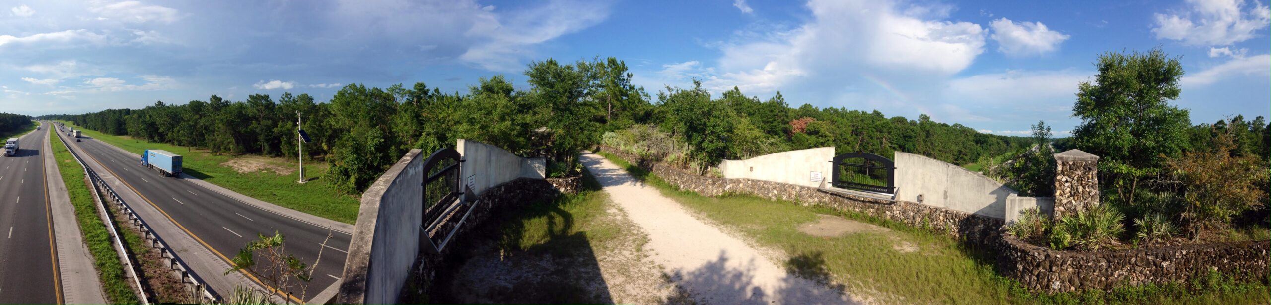 A panoramic view of a two-lane highway bordered by trees, with trucks and vehicles traveling in both directions. On the right side, there is a landscaped entrance with a stone wall and a gated pathway leading into a wooded area, under a clear blue sky with a few clouds. Santos mountain bike trail.